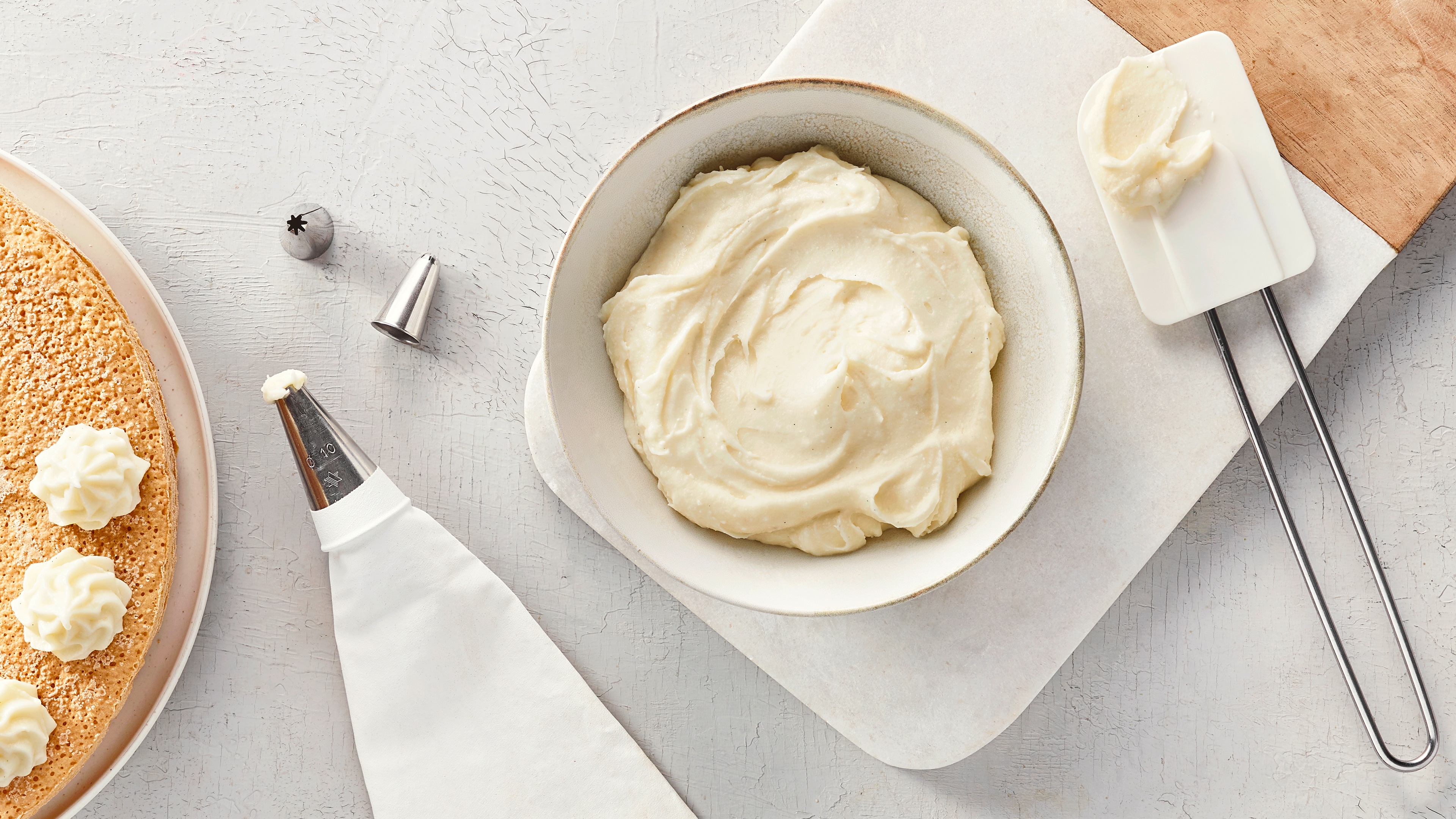 Eine Schüssel mit heller, aufgeschlagener Creme, daneben ein Spritzbeutel und eine heller Tortenboden