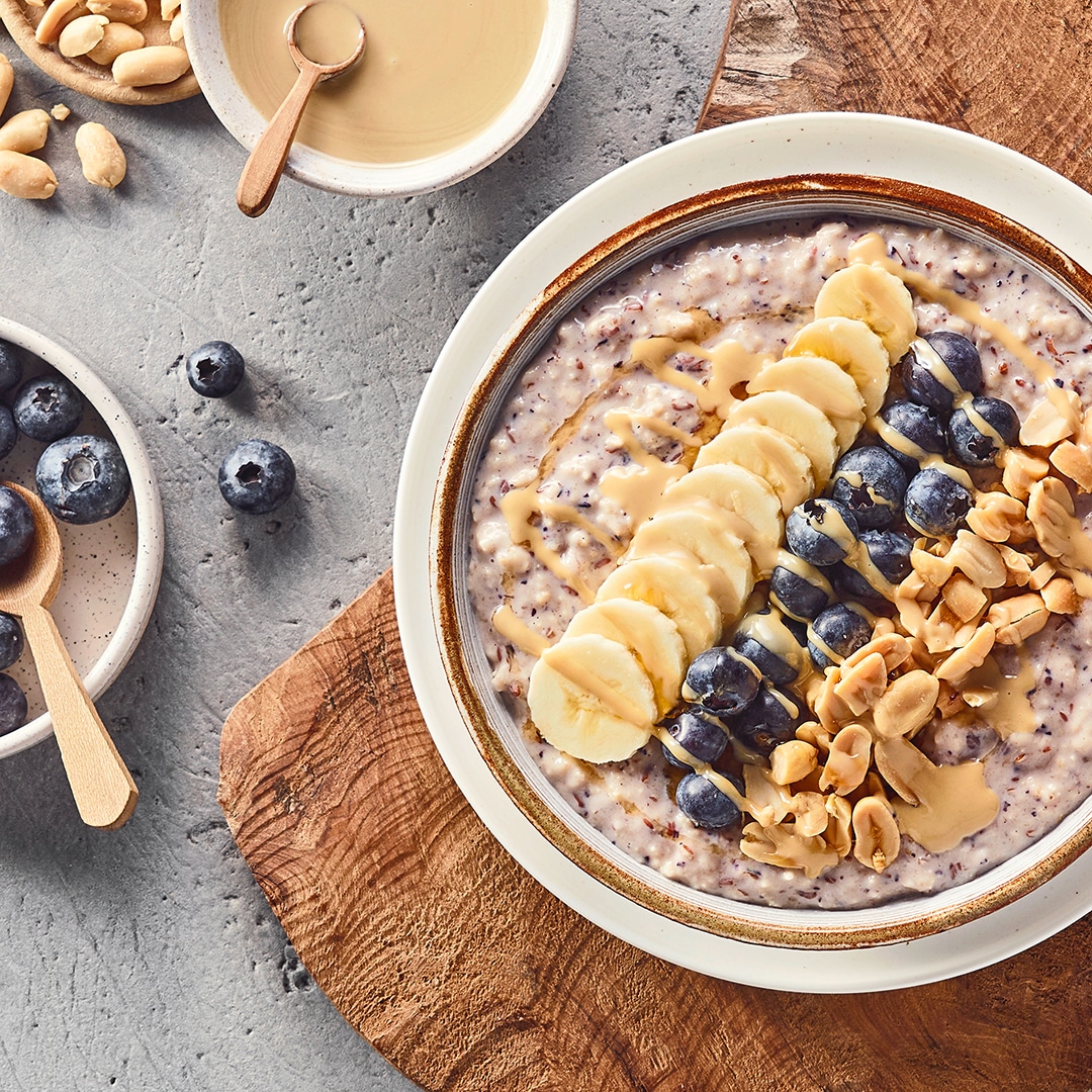 Eine Schüssel mit Haferbrei, darauf Bananenscheiben, Heidelbeeren und gehackte Erdnüsse, daneben Schalen mit Früchten.