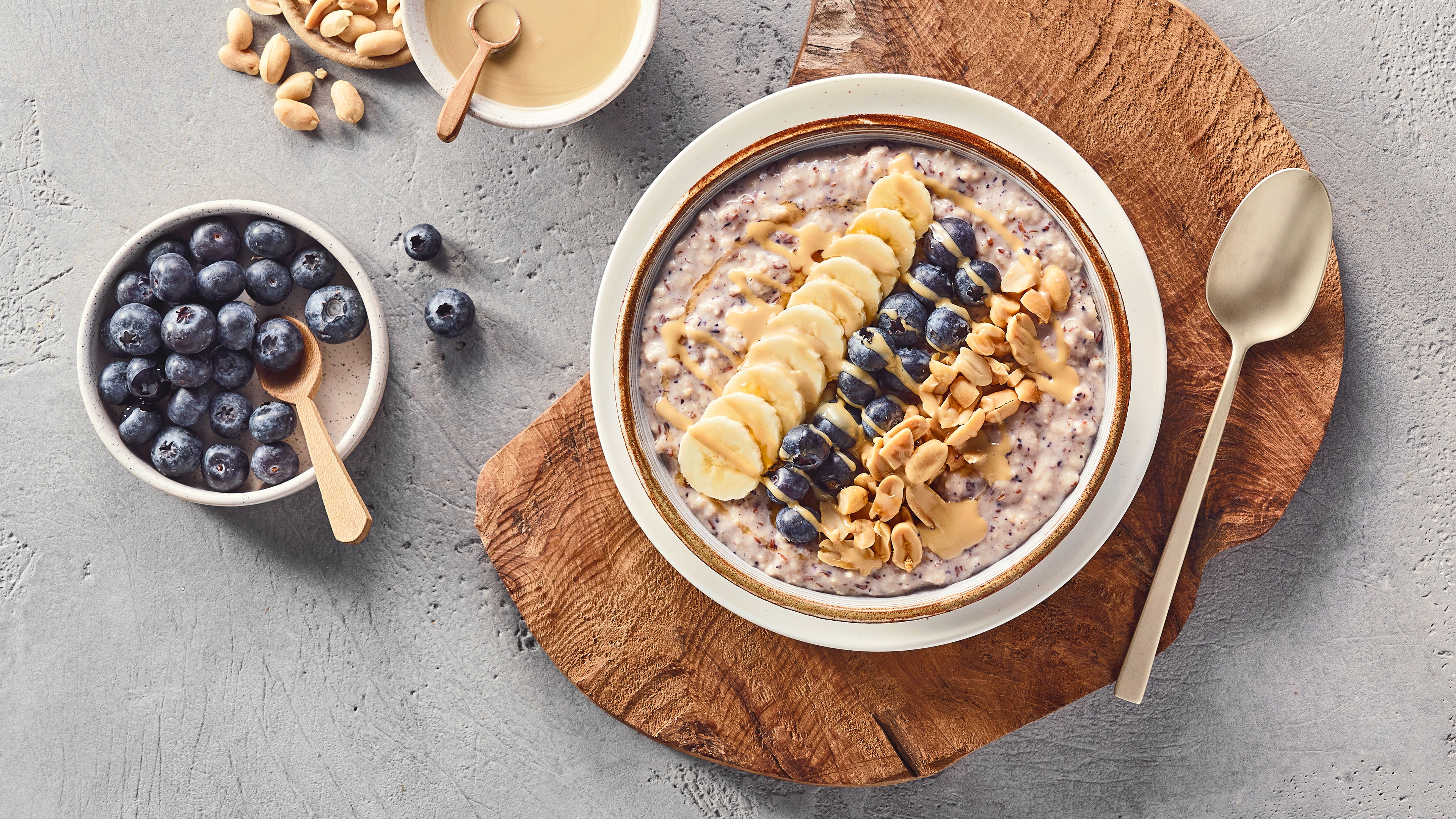 Eine Schüssel mit Haferbrei, darauf Bananenscheiben, Heidelbeeren und gehackte Erdnüsse, daneben Schalen mit Früchten.