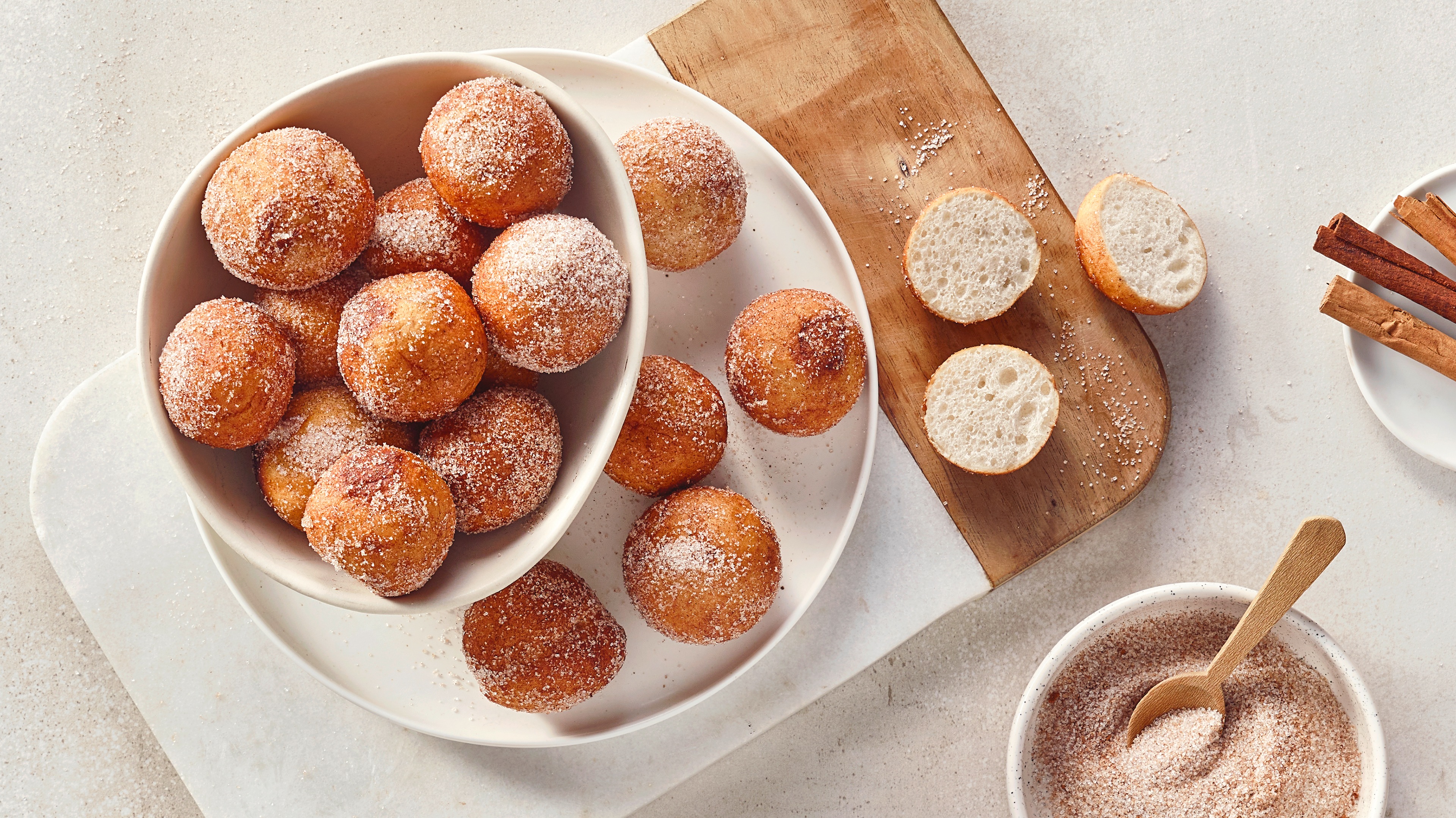 Eine Schale mit runden Teigbällchen, bestreut mit Zucker, daneben aufgeschnittene Stücke und eine Schale mit Zimt-Zucker-Mischung.
