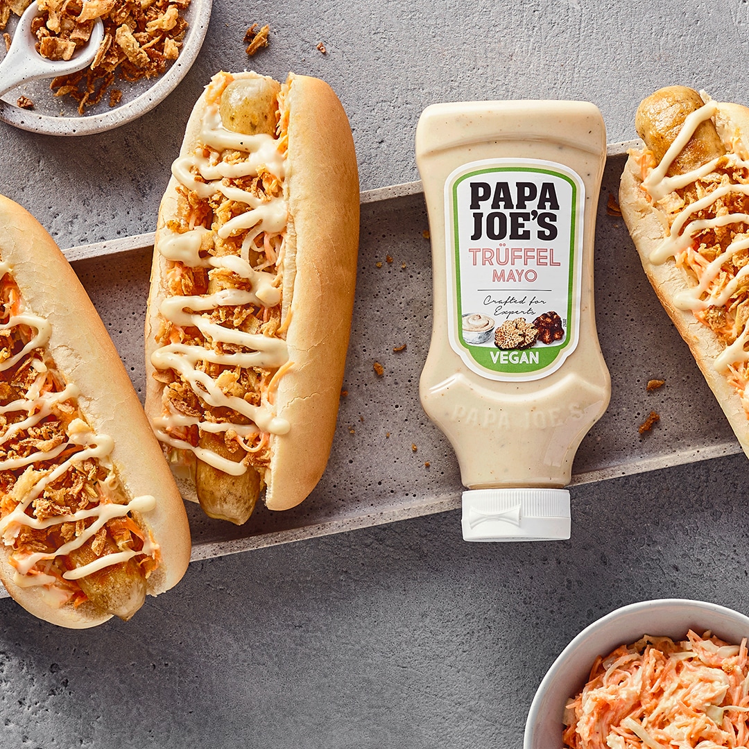 Eine rechteckige Schale mit drei Hotdogbröchten, gefüllt mit Wurst & Röstzwiebeln, daneben eine Tube PAPA JOE´S Trüffelmayo