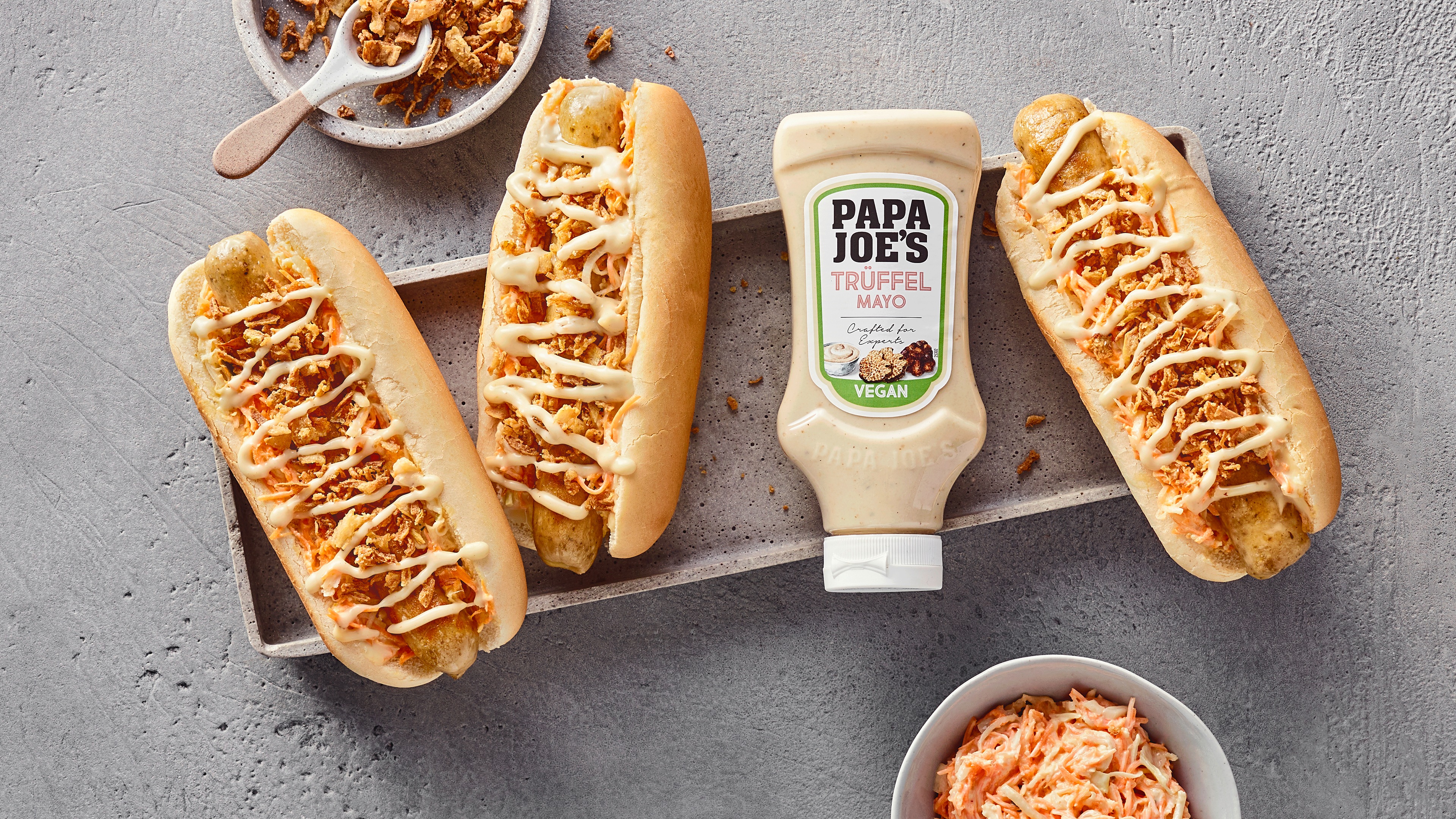 Rezept für Vegane Trüffel Hot Dogs mit Produkt Eine rechteckige Schale mit drei Hotdogbröchten, gefüllt mit Wurst & Röstzwiebeln, daneben eine Tube PAPA JOE´S Trüffelmayo