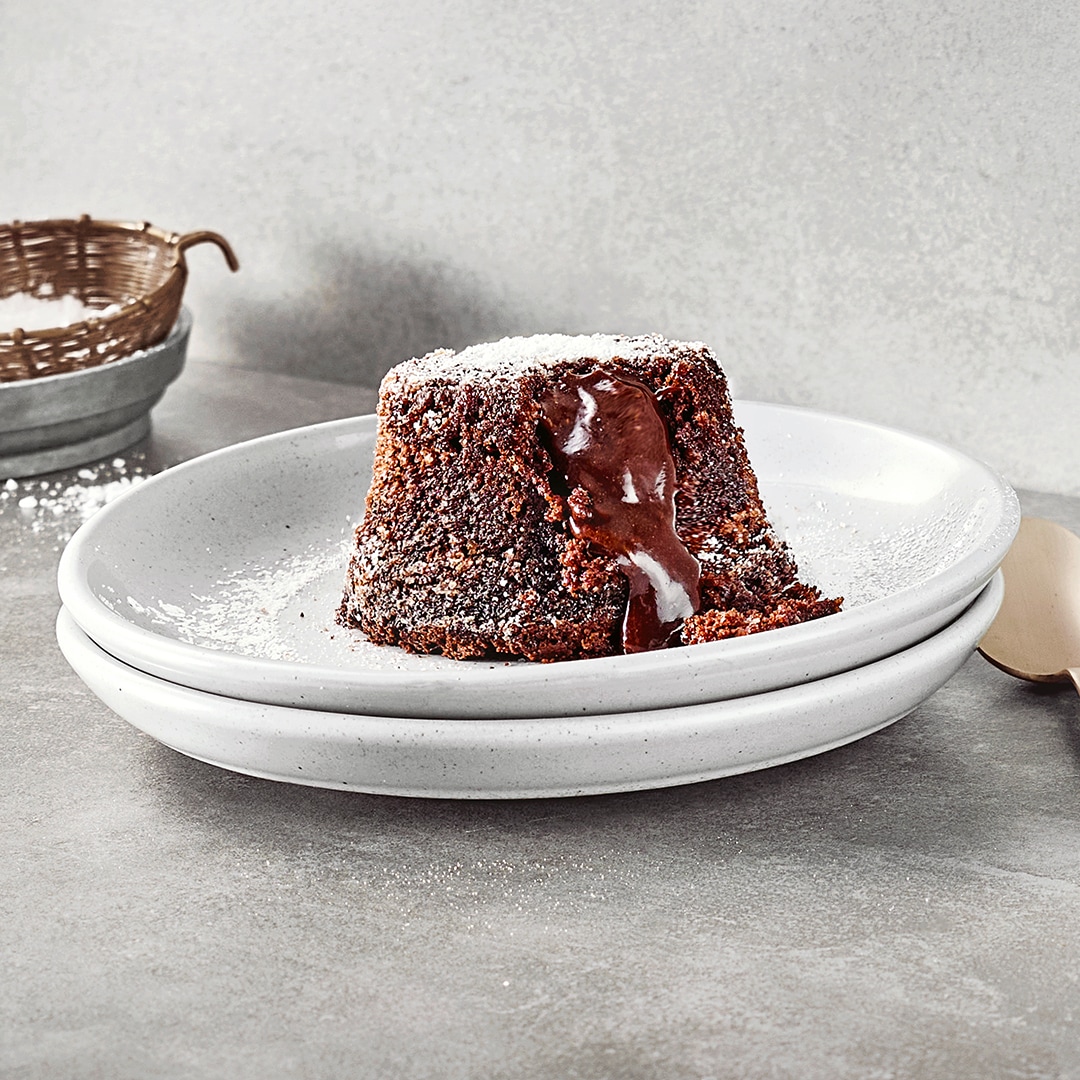 Zwei gestapelte Teller mit einem kleinen Schokoküchlein mit flüssigem Kern, bestreut mit Puderzucker. Rechts daneben liegt ein Löffel. Links oben befindet sich etwas Puderzucker zum Bestreuen.