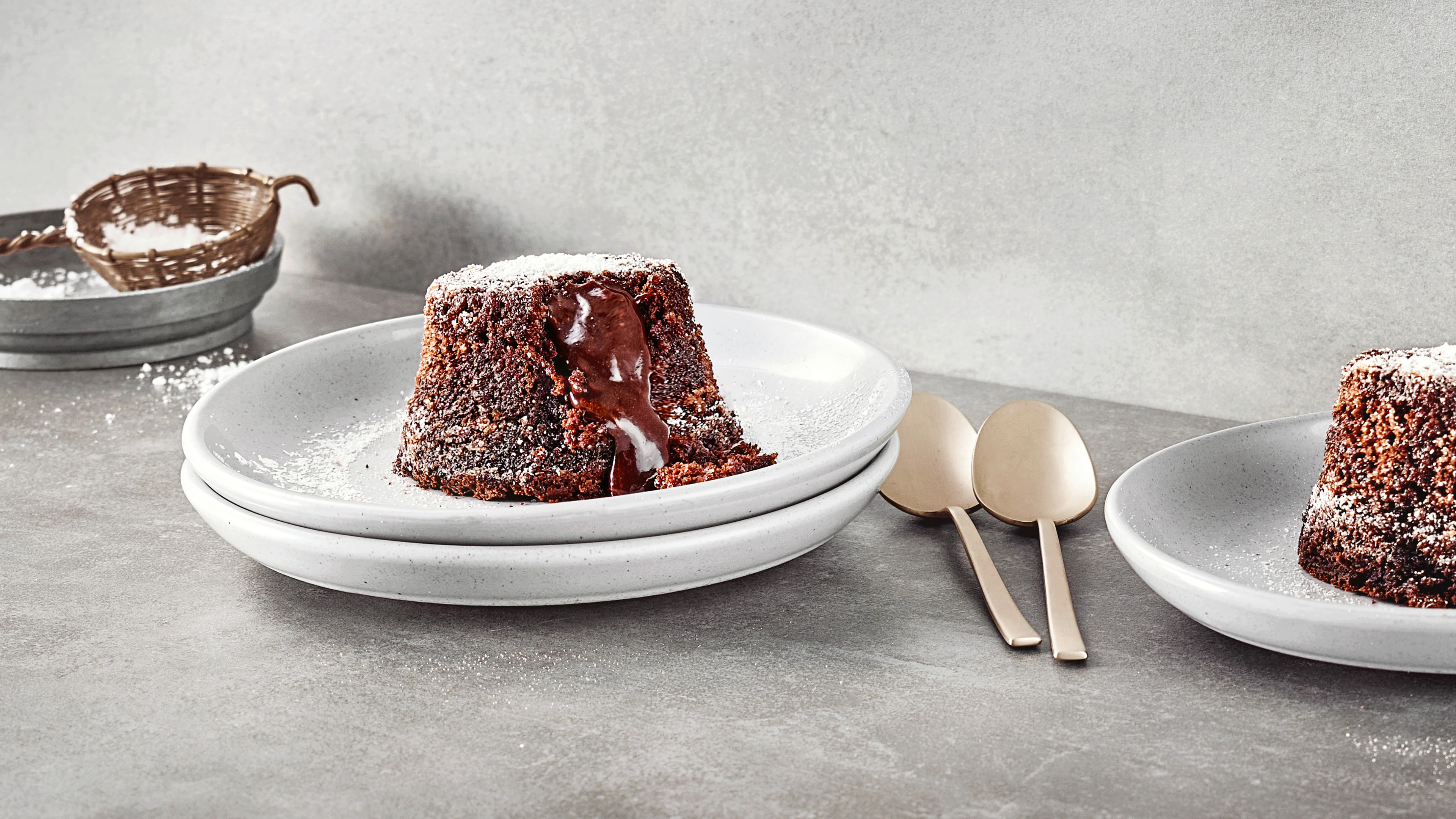 Zwei gestapelte Teller mit einem kleinen Schokoküchlein mit flüssigem Kern, bestreut mit Puderzucker. Rechts daneben liegen zwei Löffel. Links oben befindet sich etwas Puderzucker zum Bestreuen. Links unten ist abgeschnitten ein weiterer Teller mit einem Lava-Cake zu sehen.