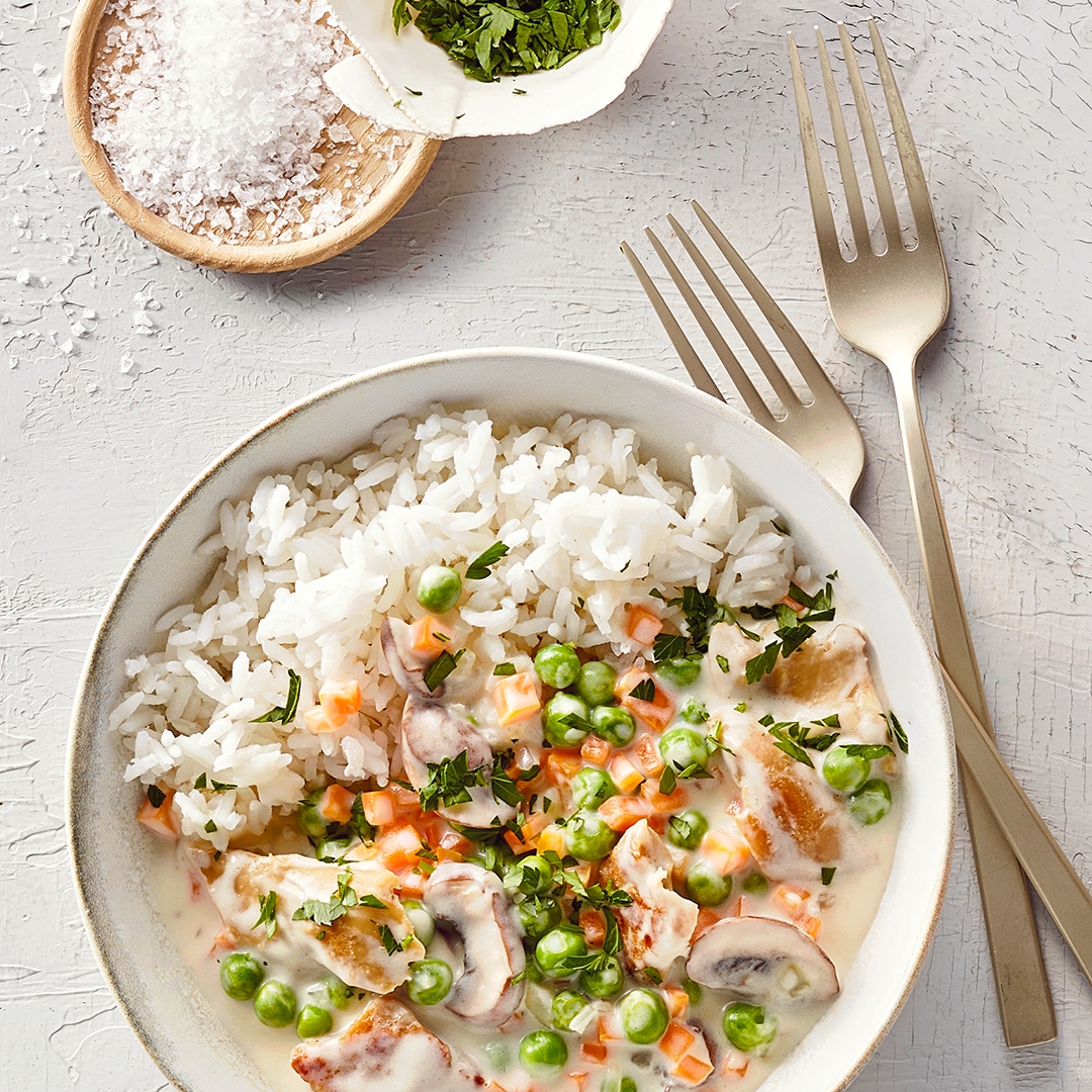 Ein Teller mit Reis, Erbsen, orangefarbenen Würfeln, Pilzscheiben & Fleischstücken in heller Soße, garniert mit Kräutern.