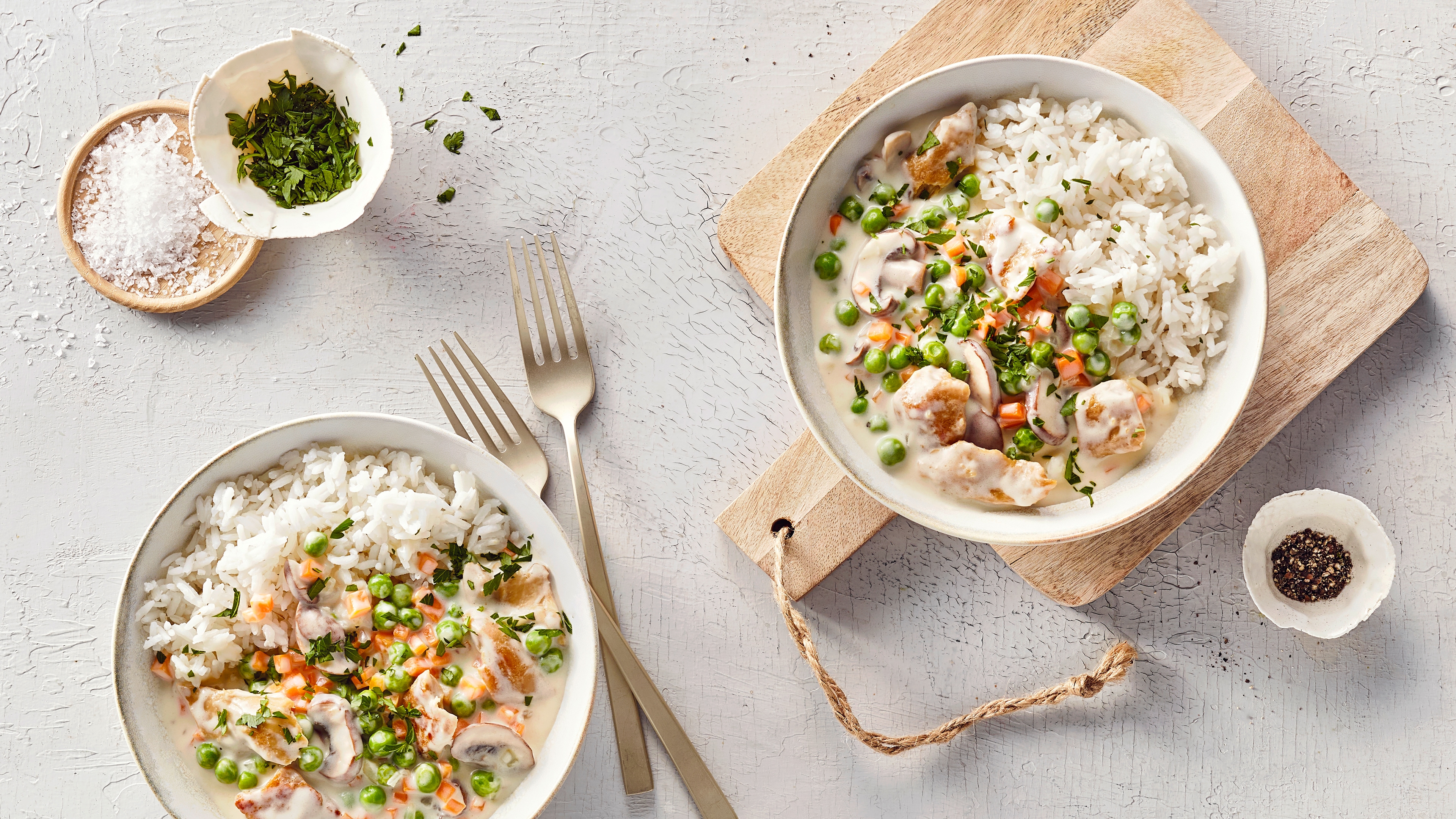 Ein Teller mit Reis, Erbsen, orangefarbenen Würfeln, Pilzscheiben & Fleischstücken in heller Soße, garniert mit Kräutern.