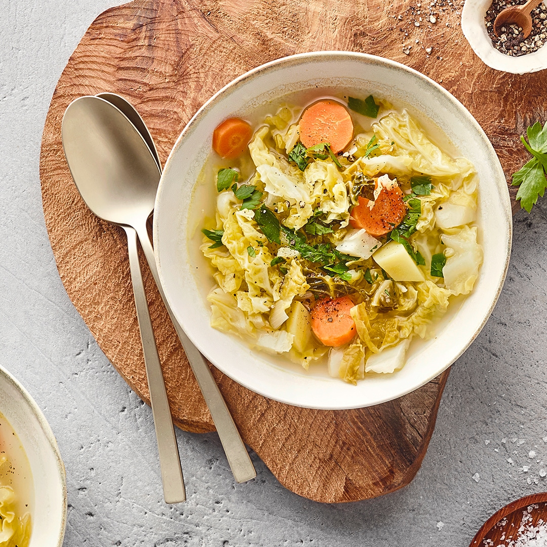 Tiefer Teller mit einem Eintopf aus Wirsing, Karotten und weiterem Gemüse, serviert auf einem Holzbrett. Daneben liegen zwei Löffel.