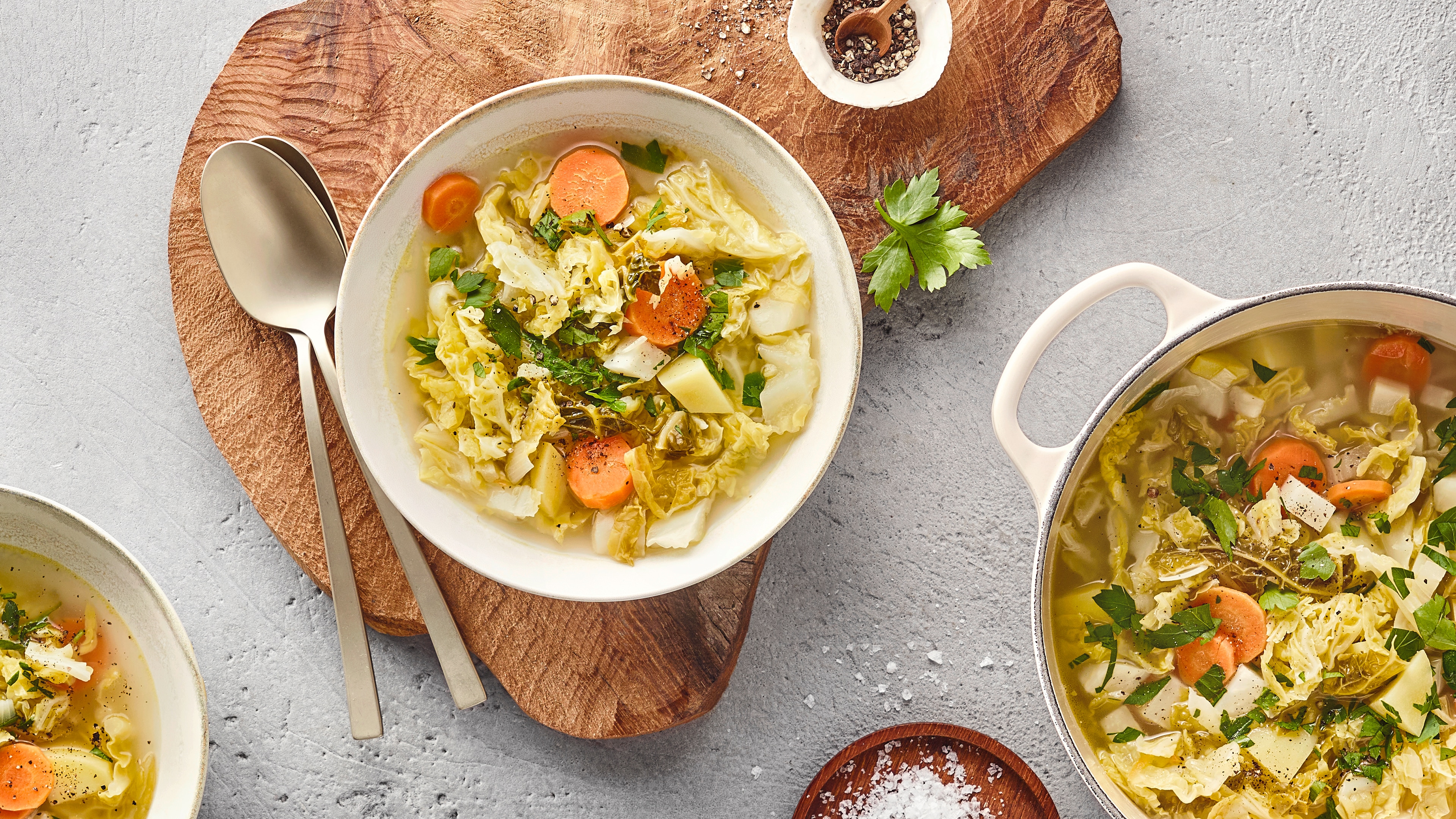 Tiefer Teller mit einem Eintopf aus Wirsing, Karotten und weiterem Gemüse, serviert auf einem Holzbrett. Daneben liegen zwei Löffel. Rechts unten steht ein größerer Topf mit weiterem Eintopf.