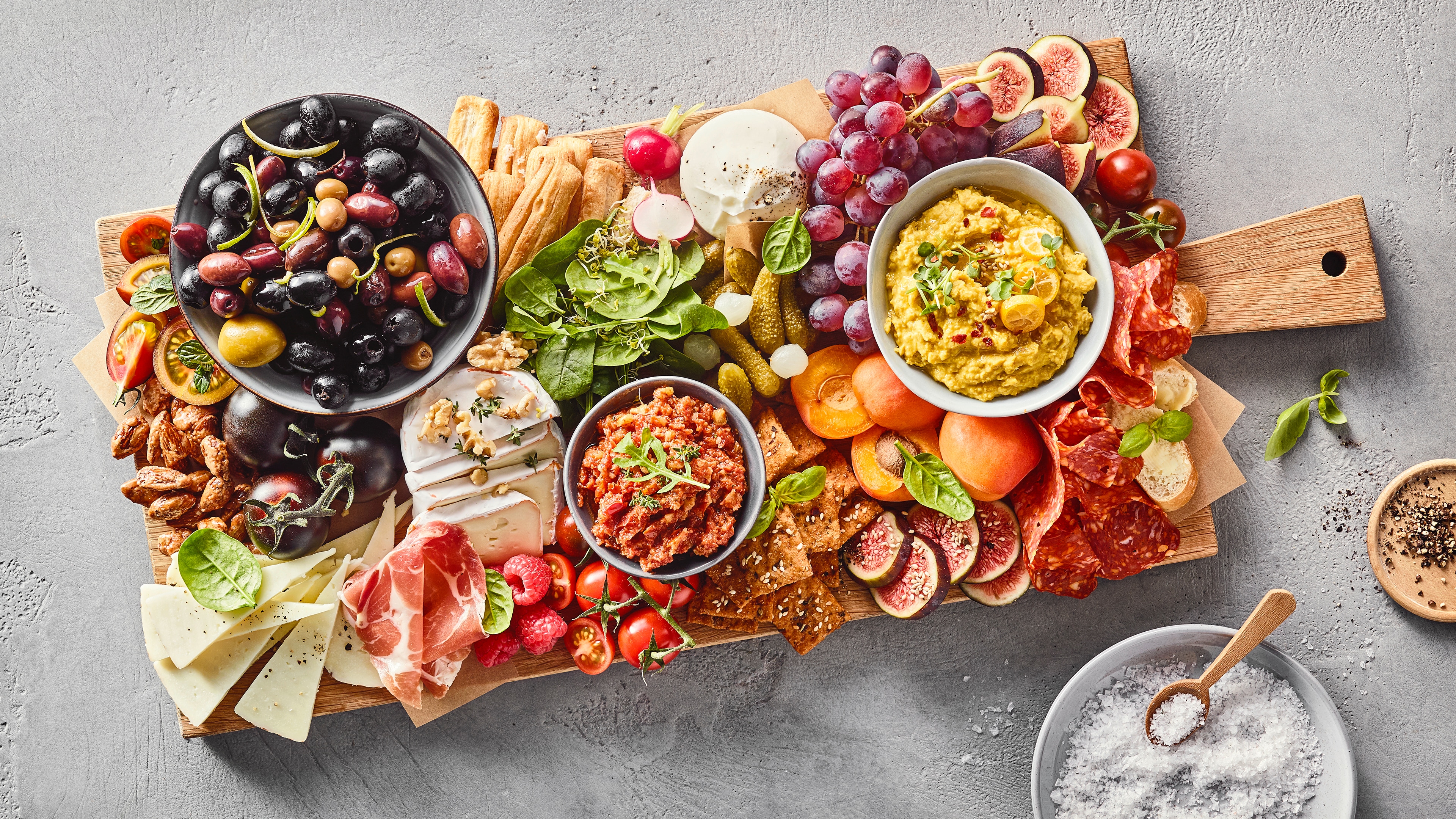 Ein Brett mit schwarzen und grünen Oliven, Trauben, Feigen, Käse, Schinken, Salami, Crackern, grünen Blättern und zwei Schalen mit Dips.