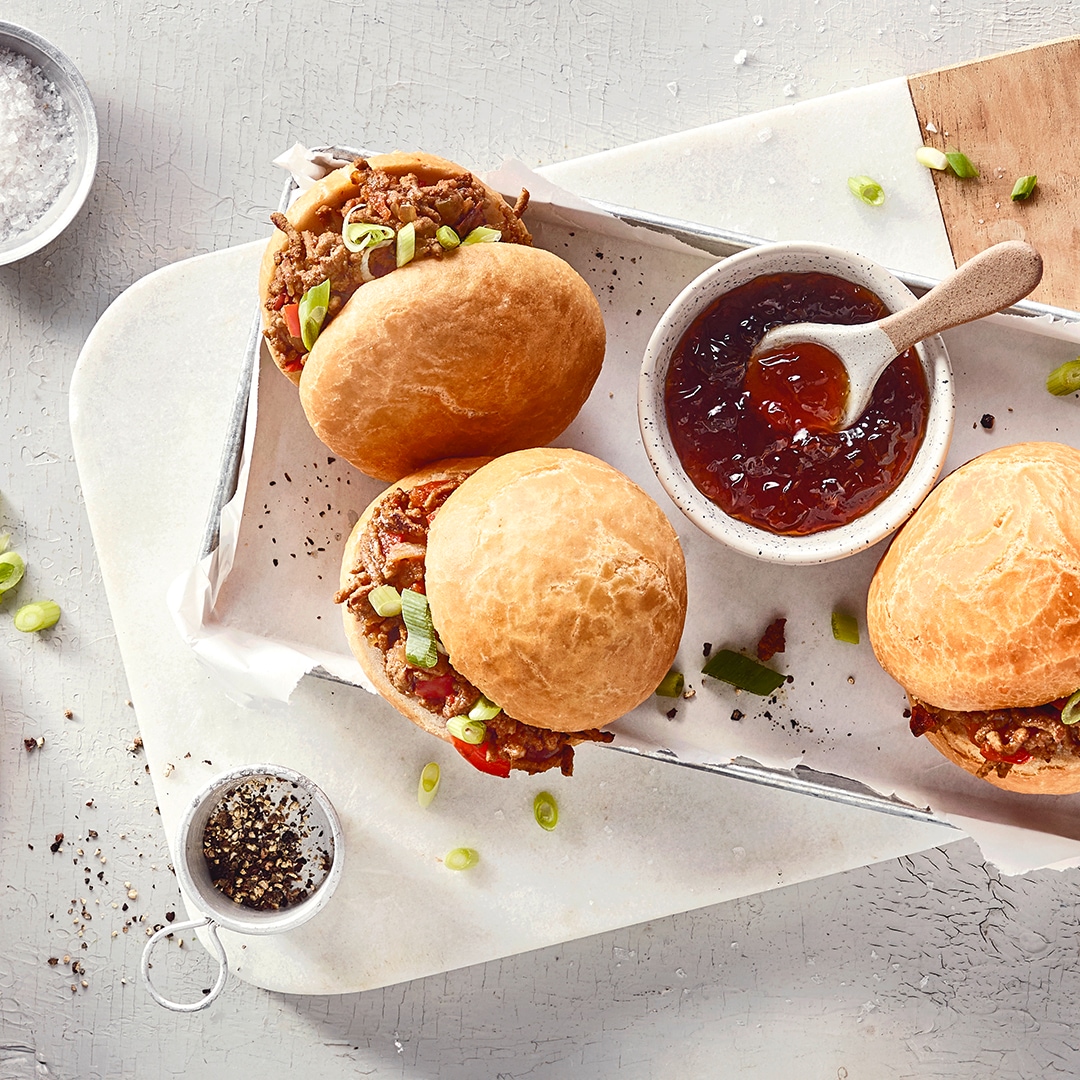 Ein Brett mit runden Brötchen, gefüllt mit braunen Stücken und grünen Ringen, daneben eine Schale mit roter, glänzender Soße.