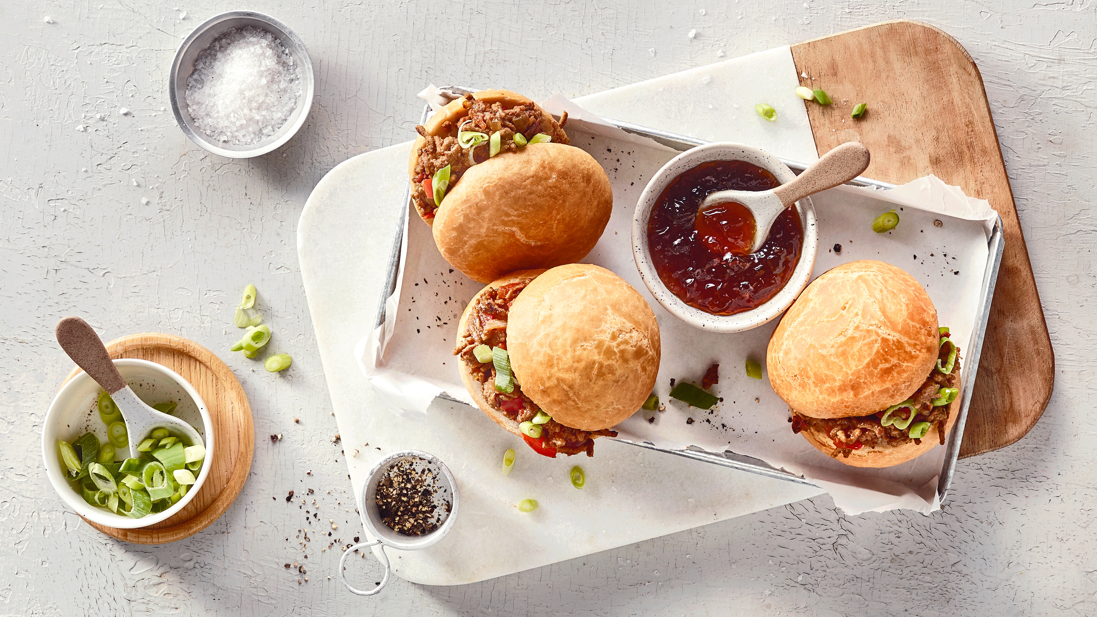 Ein Brett mit runden Brötchen, gefüllt mit braunen Stücken und grünen Ringen, daneben eine Schale mit roter, glänzender Soße.