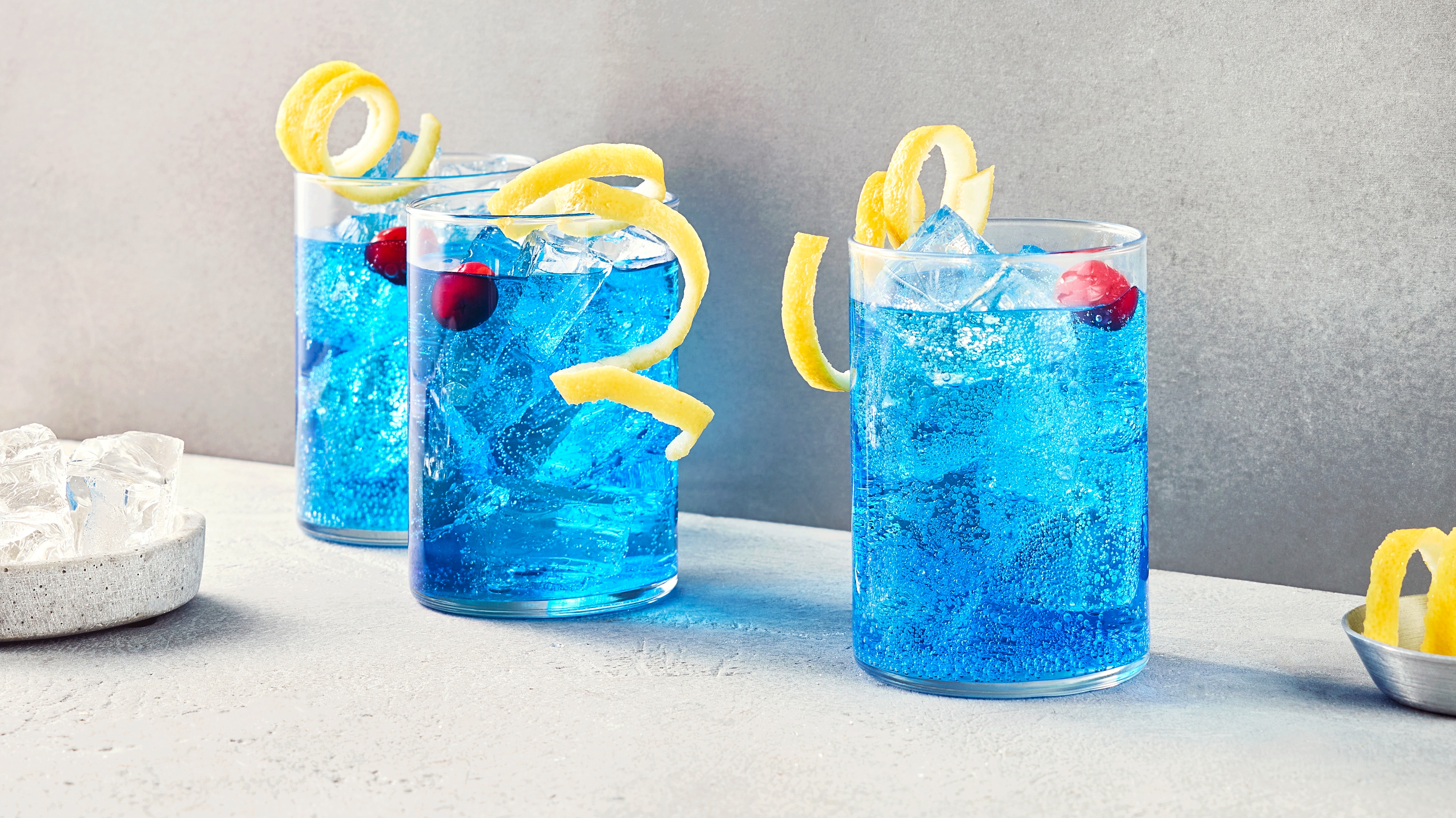 Gläser mit blauer Flüssigkeit, Eiswürfel, Kirsche und Zitronenzeste