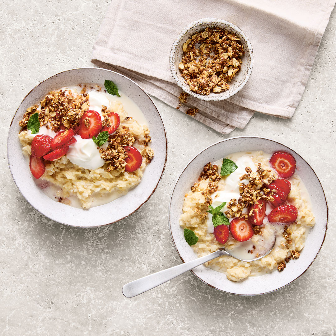 Zwei Schälchen mit Milchreis, Erdbeerscheiben und Granola. Daneben ein Schälchen Granola