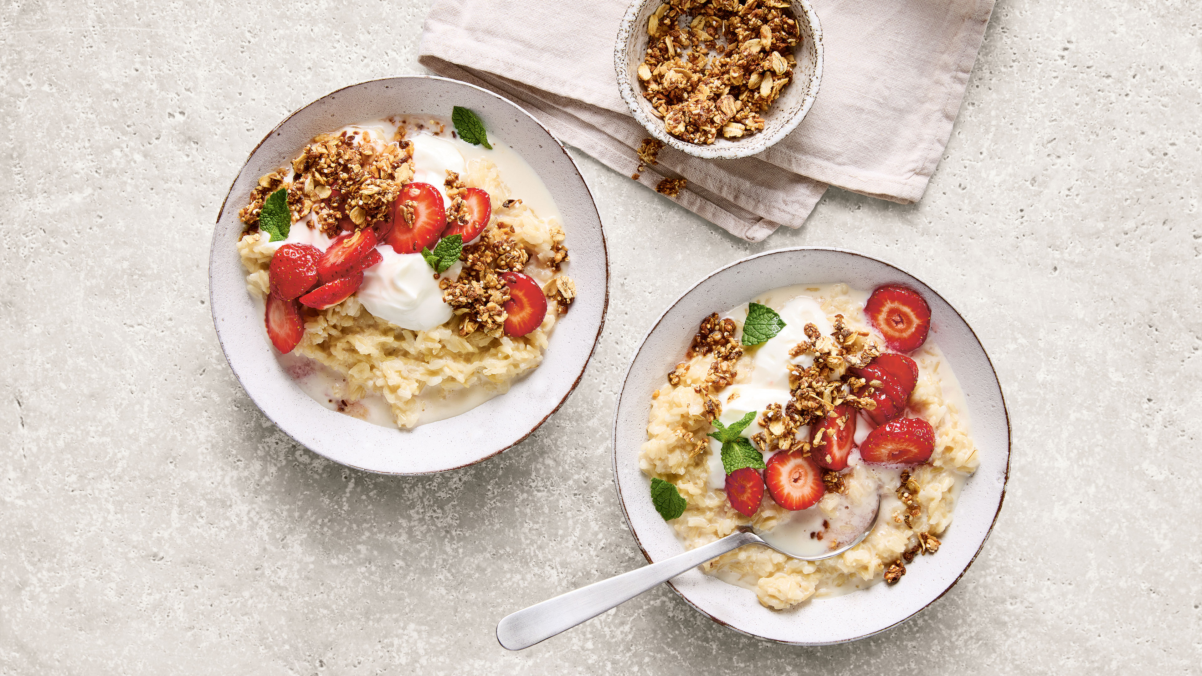 Zwei Schälchen mit Milchreis, Erdbeerscheiben und Granola. Daneben ein Schälchen Granola