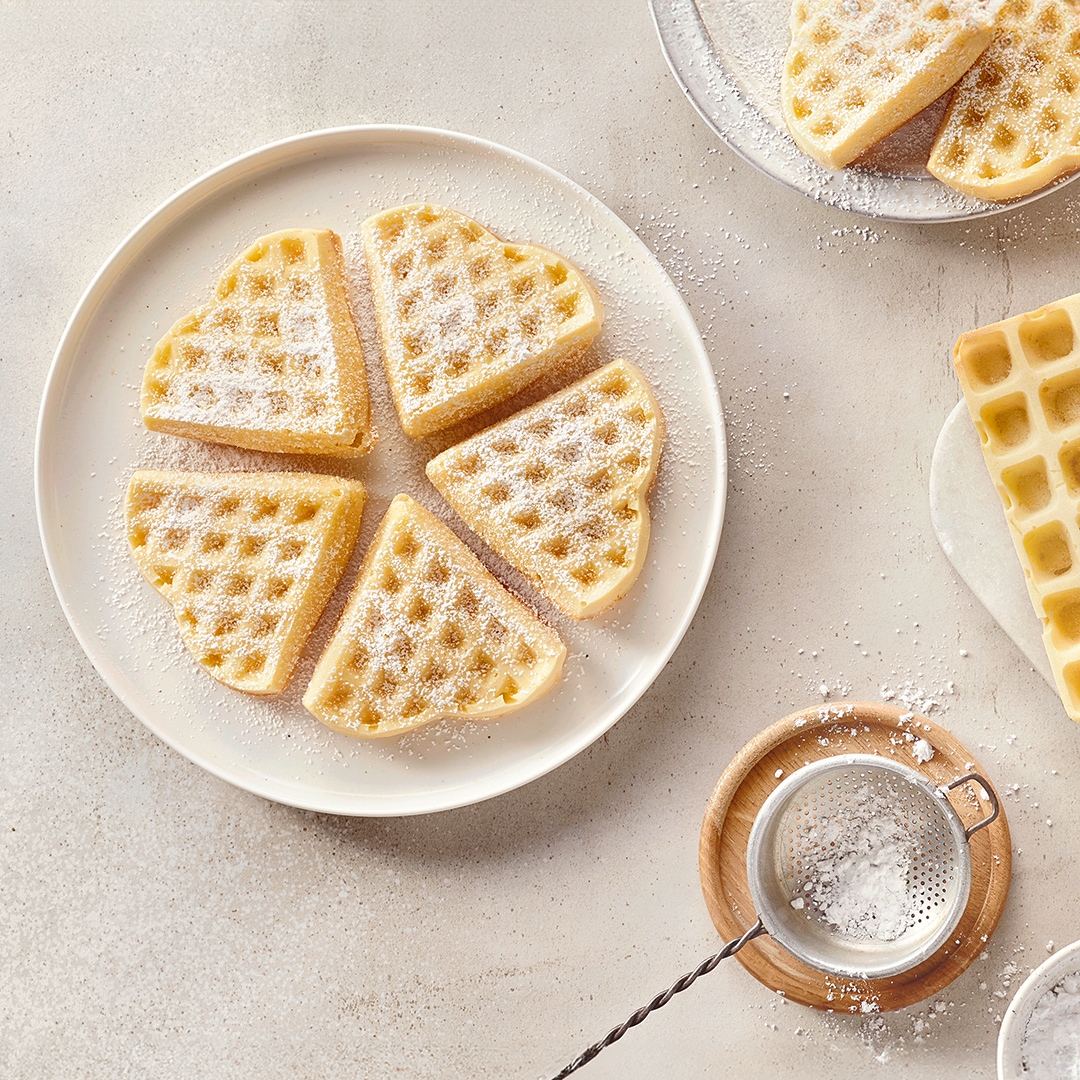 Ein Teller mit hellen Waffelstücken in Herzform, bestreut mit weißem Puderzucker, daneben ein Sieb und weitere Waffeln.