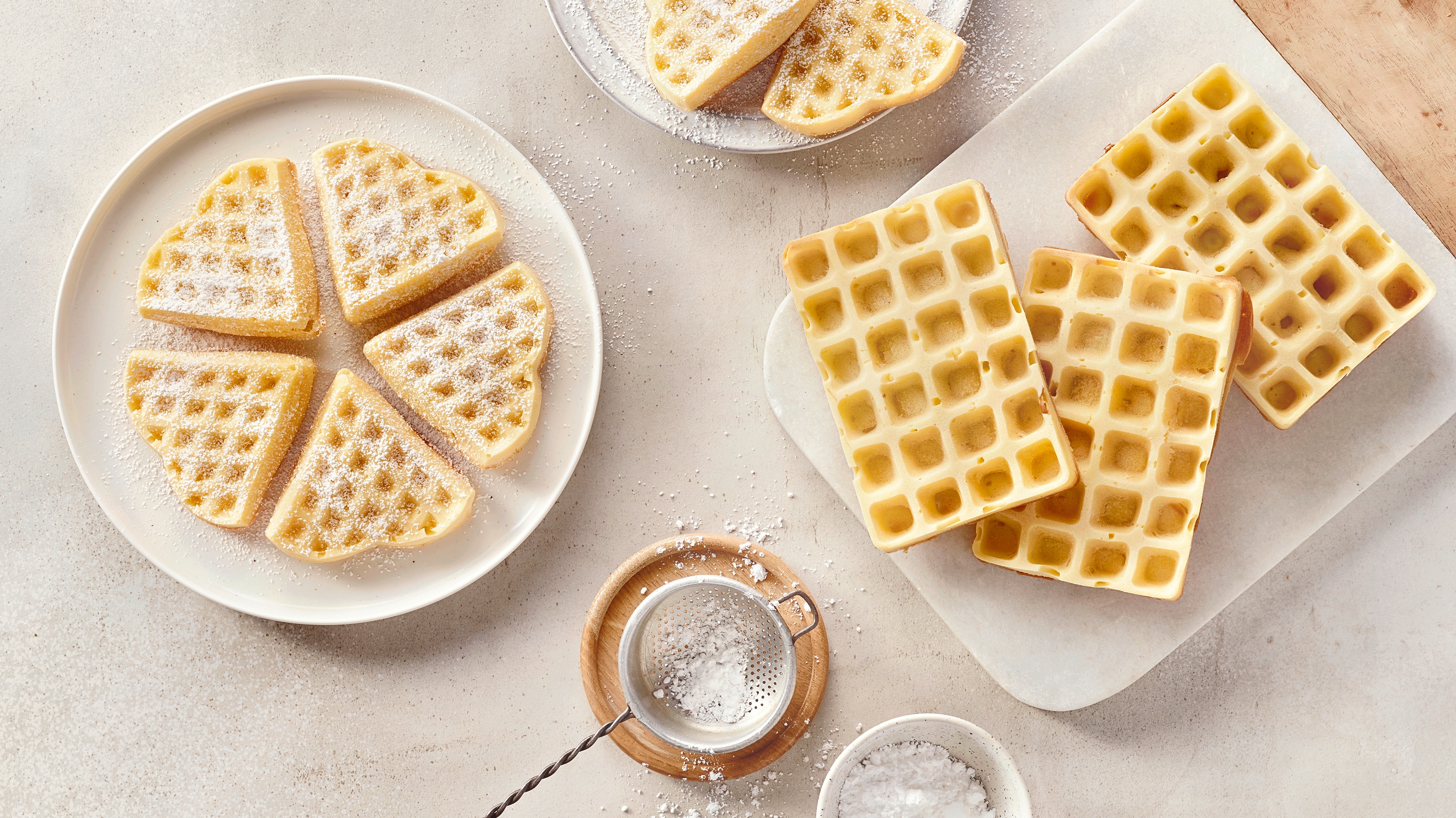 Ein Teller mit hellen Waffelstücken in Herzform, bestreut mit weißem Puderzucker, daneben ein Sieb und weitere Waffeln.