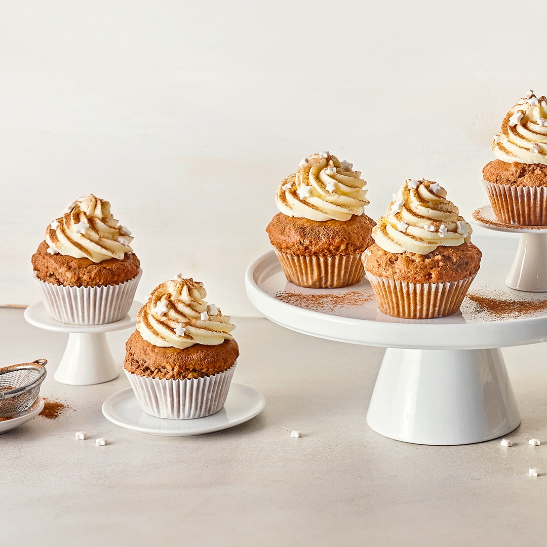 Mittelbraune Muffins, getoppt mit einer aufgespritzter Creme, garniert mit weißen Zuckersternchen, auf Keramiketageren