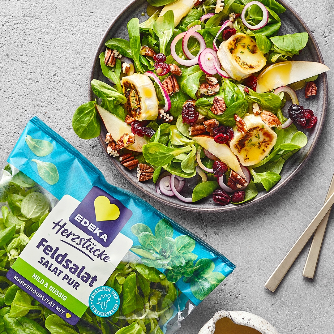 Salatteller mit Feldsalat, gebratenen Bananenstücken, Apfelstücken, Cranberries, Zwiebeln und Pekannüssen. Links unten EDEKA Herzstücke Feldsalat.