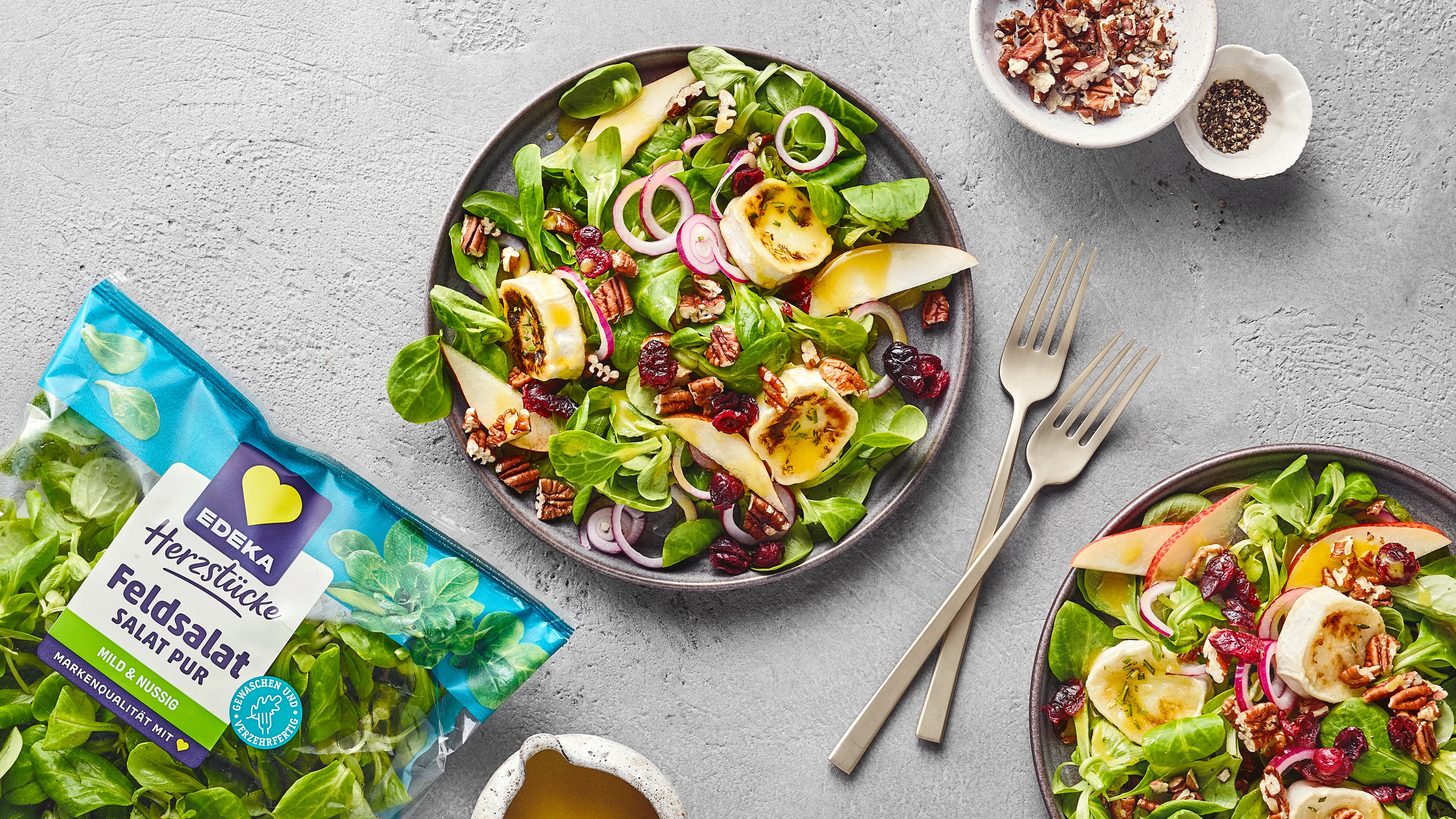 Zwei Salatteller mit Feldsalat, gebratenen Bananenstücken, Apfelstücken, Cranberries, Zwiebeln und Pekannüssen. Links unten EDEKA Herzstücke Feldsalat.