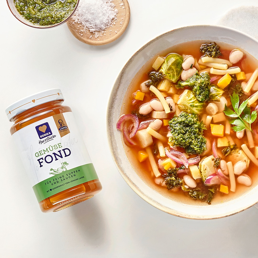 Gemüsesuppe mit weißen Bohnen, Nudeln und Pesto in einer Schale, daneben EDEKA Herzstücke Gemüsefond, Pesto und Salz.