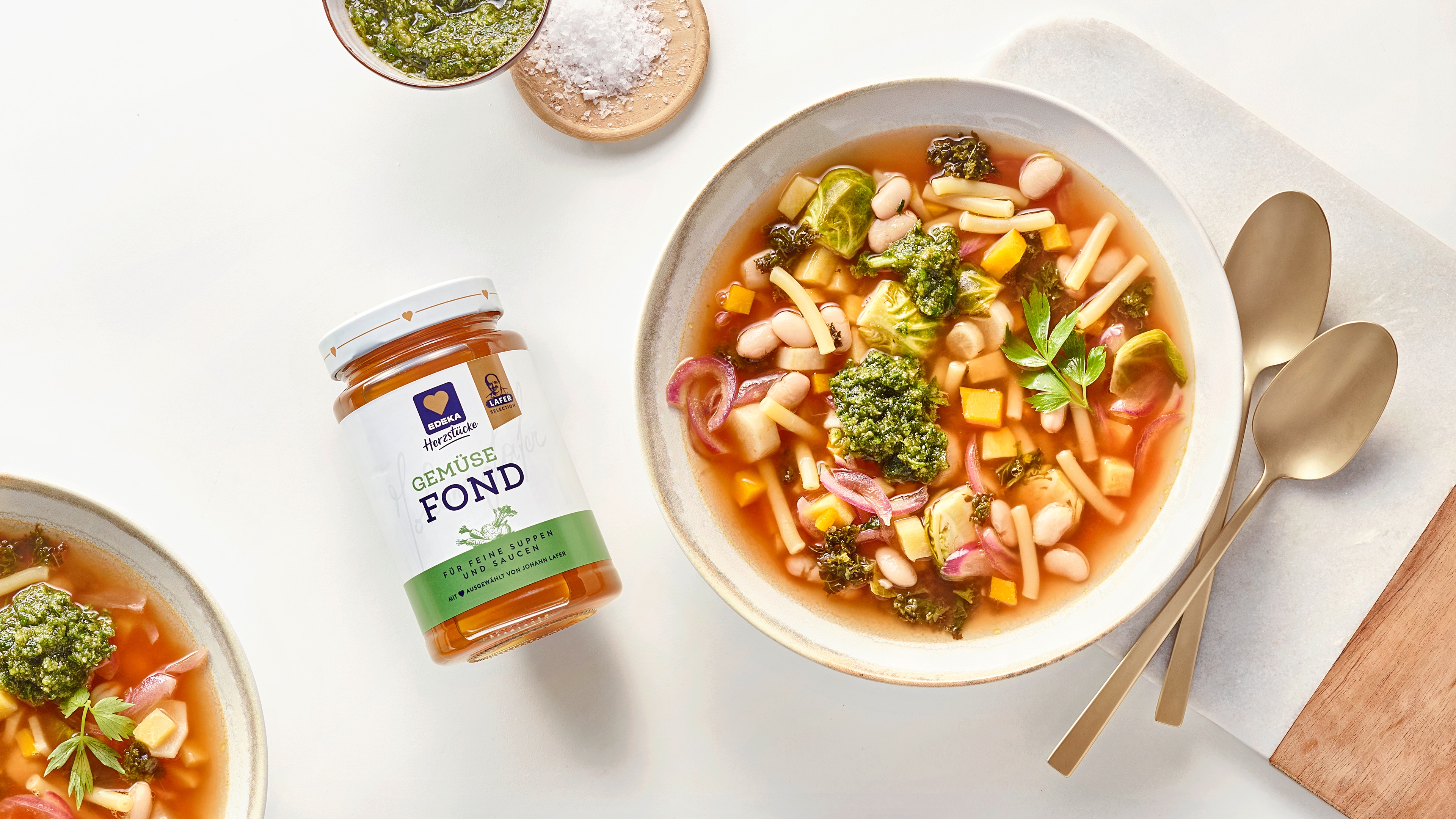 Gemüsesuppe mit weißen Bohnen, Nudeln und Pesto in einer Schale, daneben EDEKA Herzstücke Gemüsefond, zwei Löffel sowie Pesto und Salz.