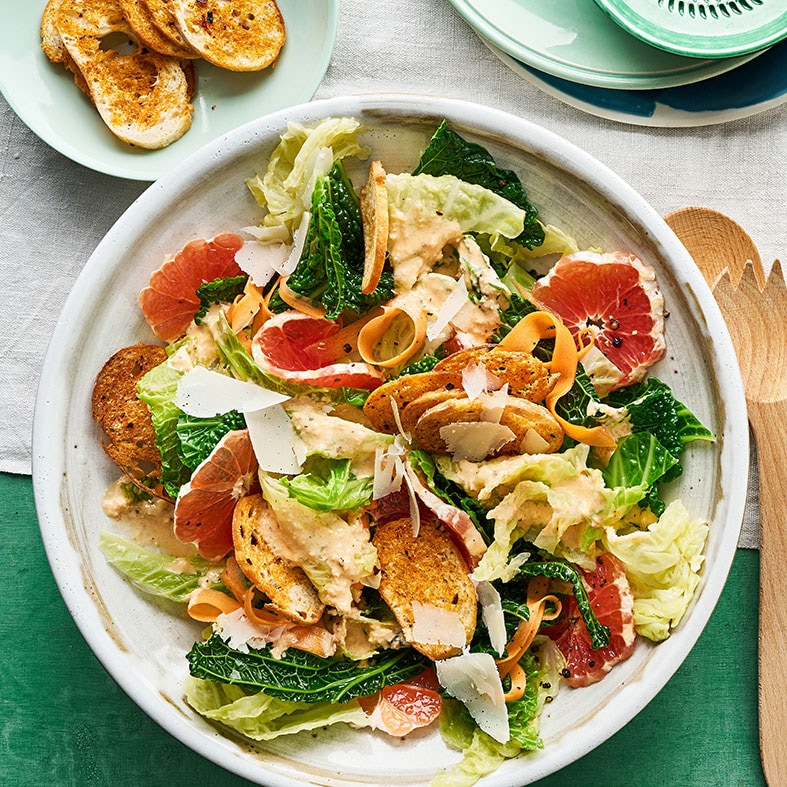 Ein Teller mit grünen Salatblättern, orange Karottenstreifen, roten Zitrusstücken, Brotscheiben und weißen Käsespänen.