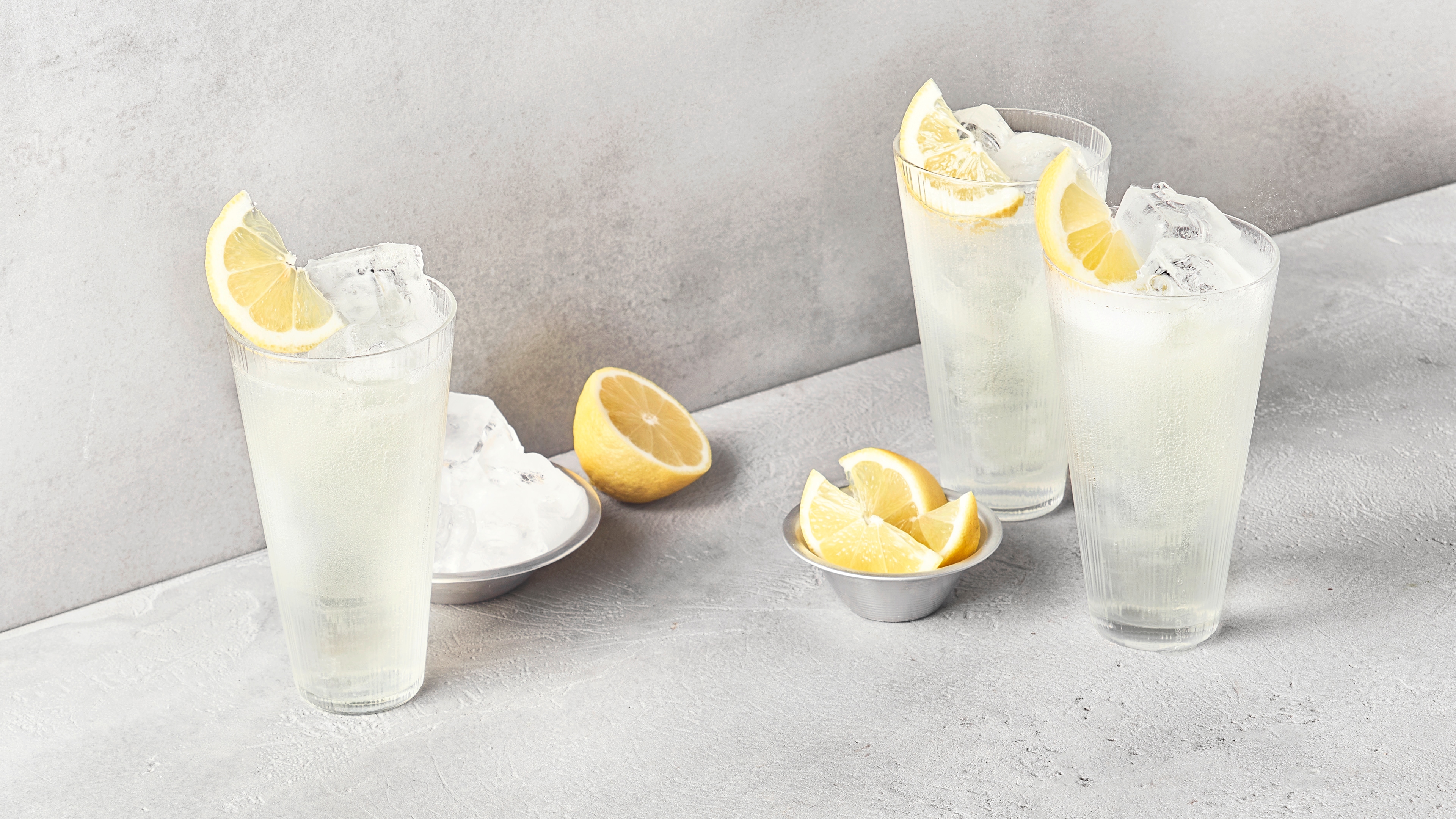 Zwei Gläser, gefüllt mit klarer Flüssigkeit, Eiswürfeln und gelben Zitronenspalten sowie einer Schale mit weiteren Zitronenstücken.
