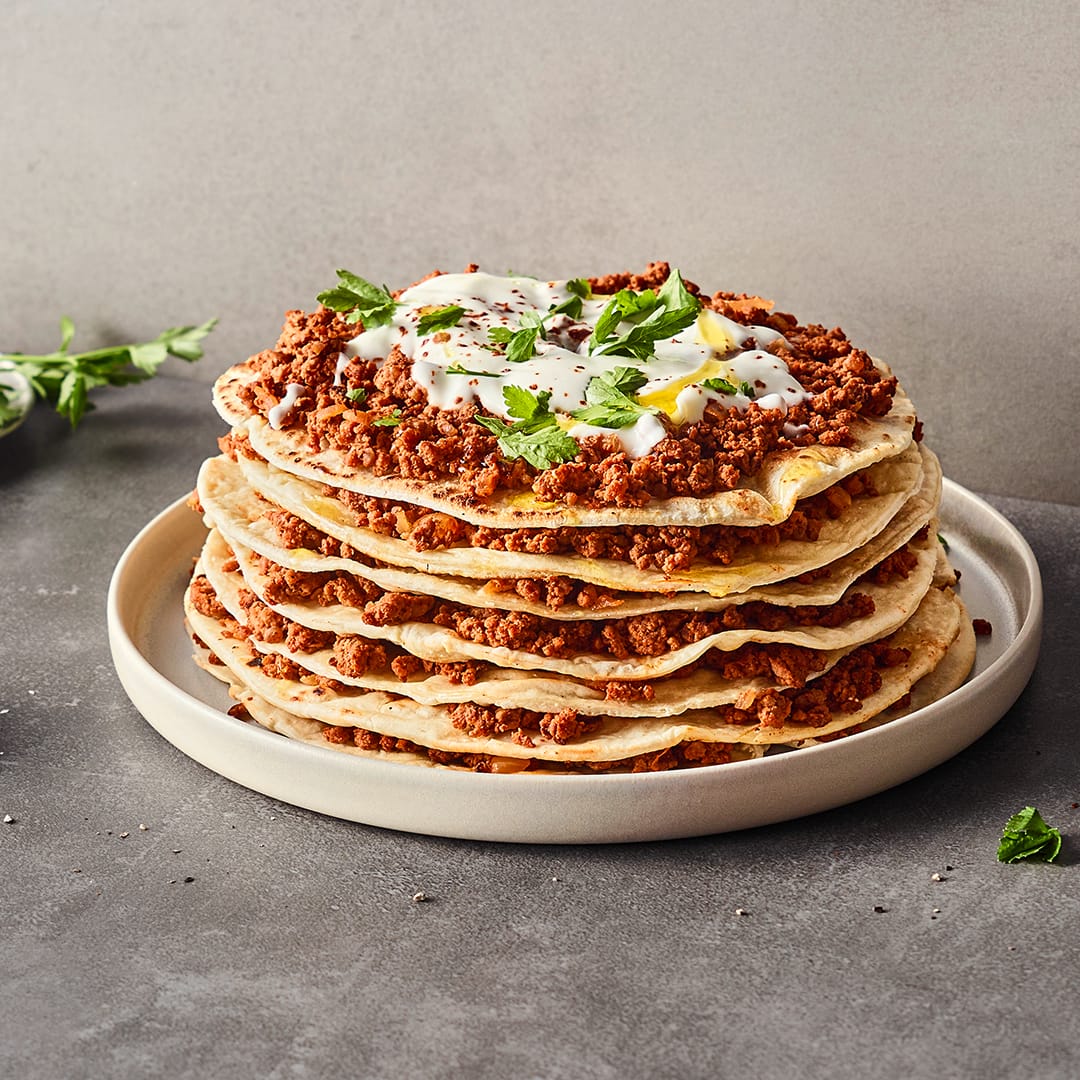 Auf einem Teller ein Lasagne-Turm aus geschichten dünnen Fladenbroten und Hackfleisch, getoppt mit leichter Joghurtsoße