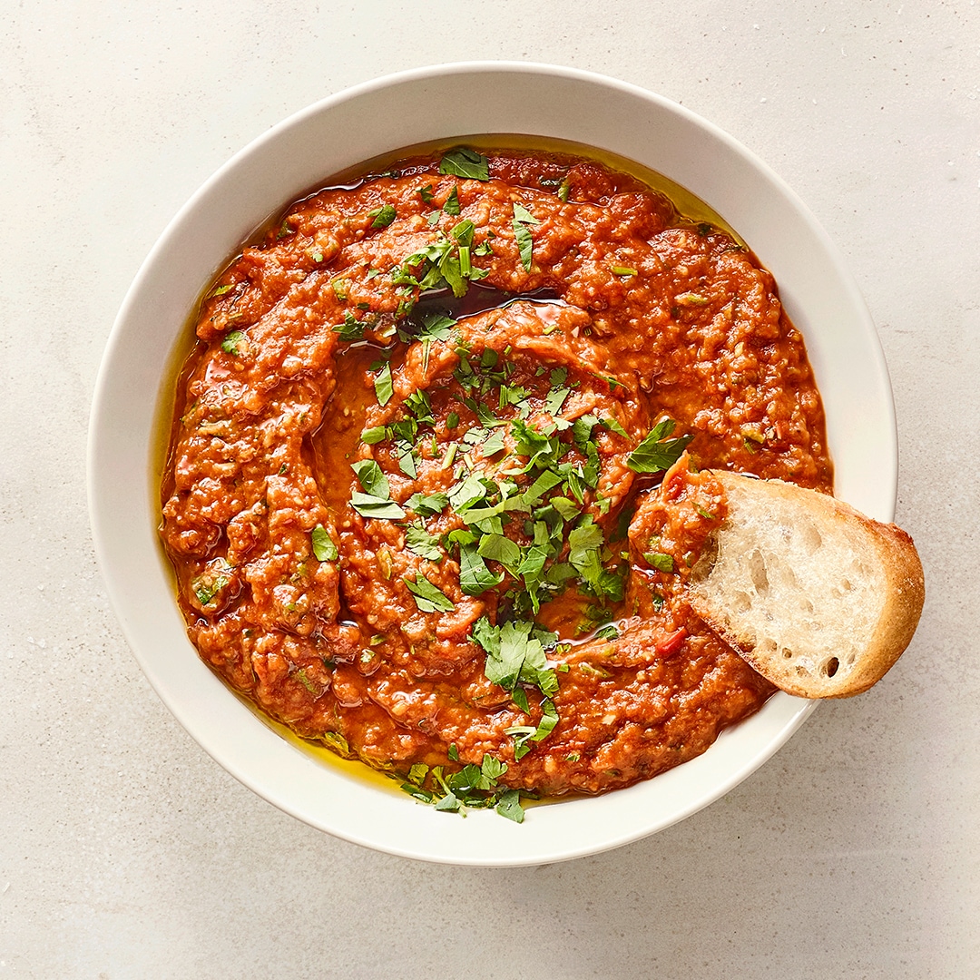 Eine Schüssel mit roter stückiger Paste, beträufelt mit Öl, bestreut mit frischen Kräutern und einem Stück Weißbrot am Rand.