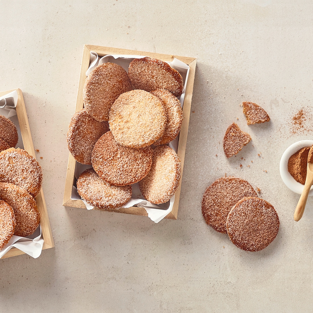 Eine Holzschale mit runden Keksen, mit Zucker bestreut, daneben zerbrochene Stücke und eine kleine Schale mit Zimt