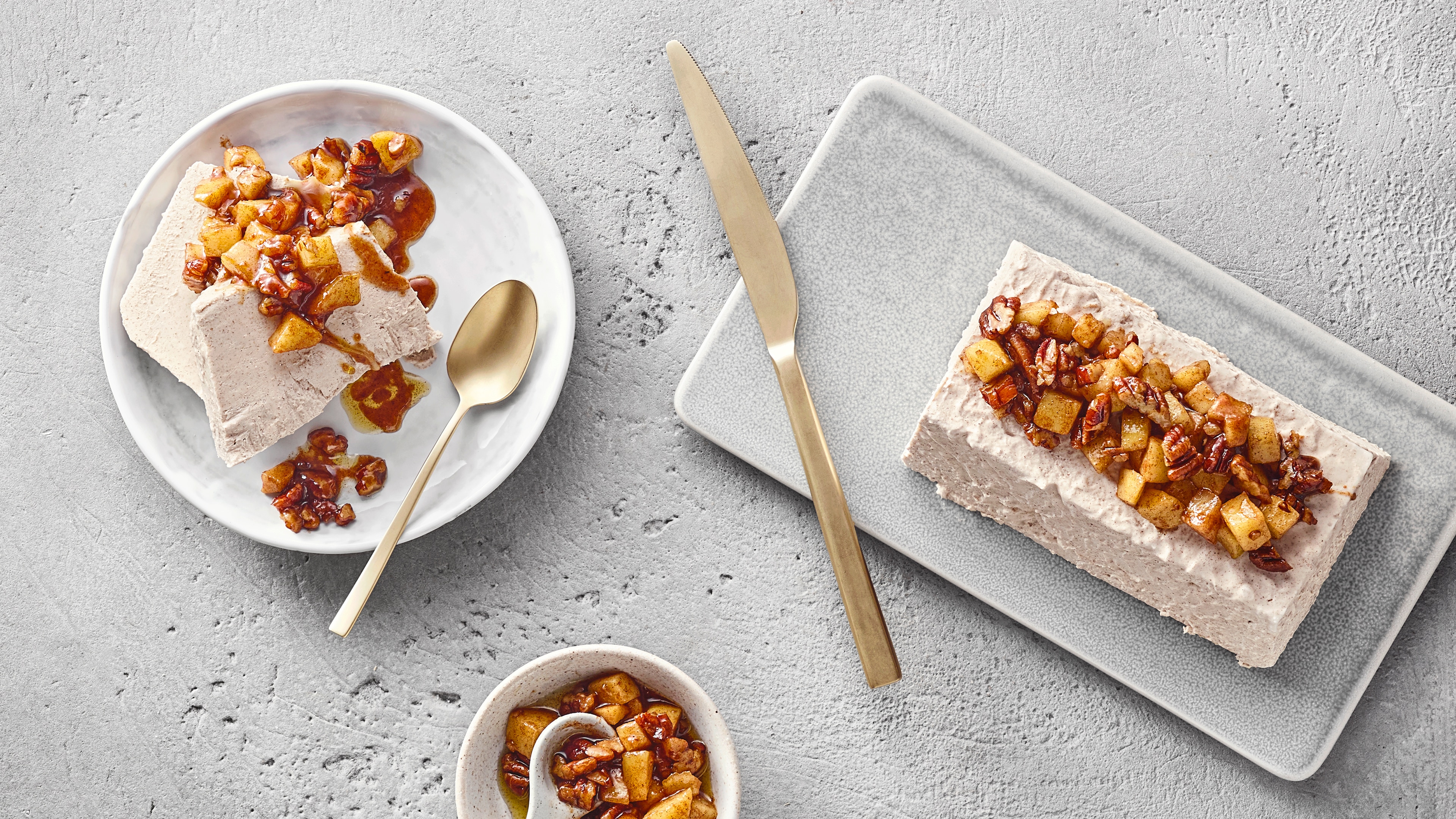 Helle cremige Scheiben mit Soße mit Nussstücken auf hellem Teller. Daneben Servierplatte mit kastenförmiger Creme und Garnitur.