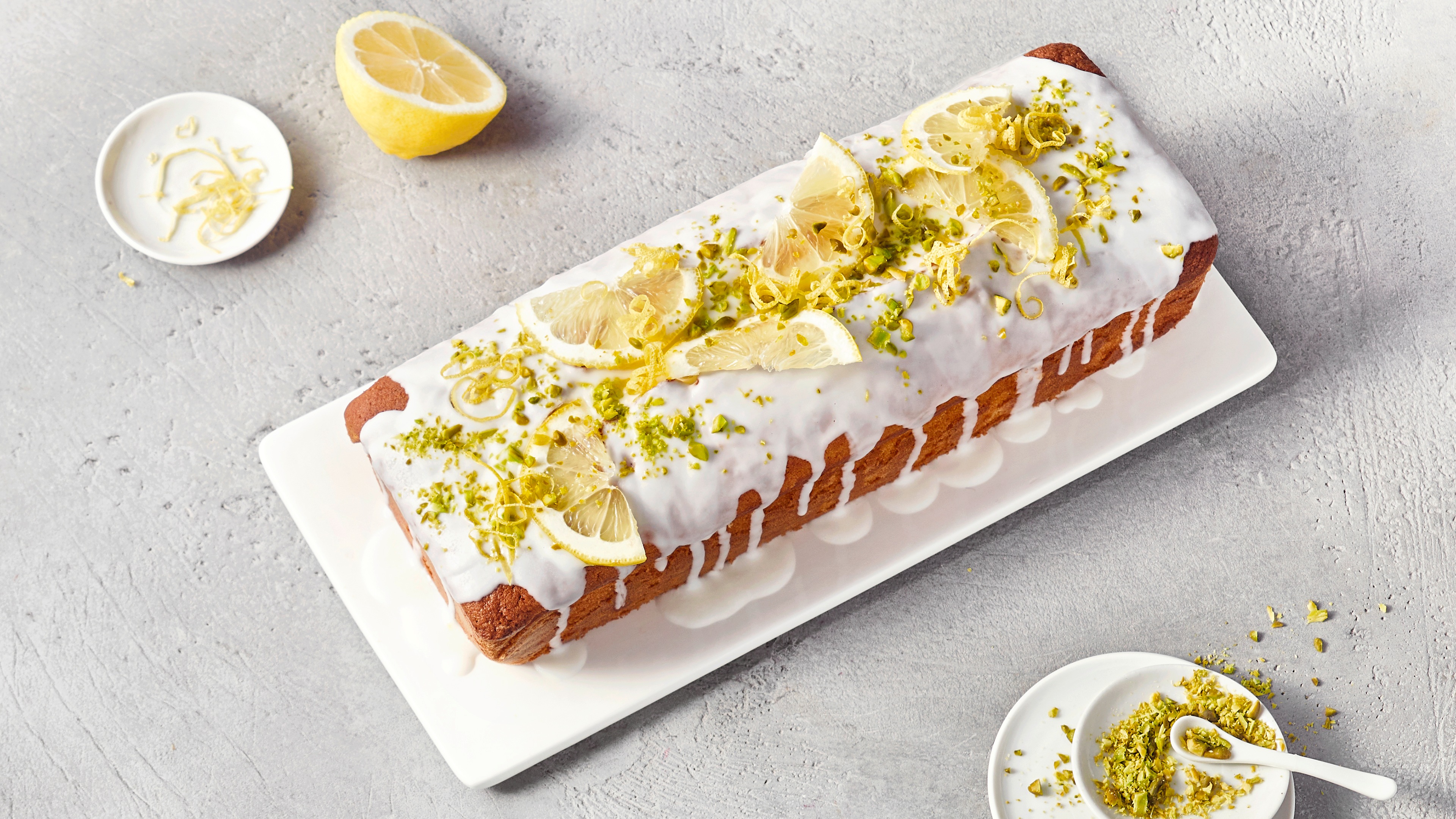 Ein Kuchen mit weißem Guss, belegt mit Zitronenscheiben und grünem Abrieb auf einer weißen Platte.