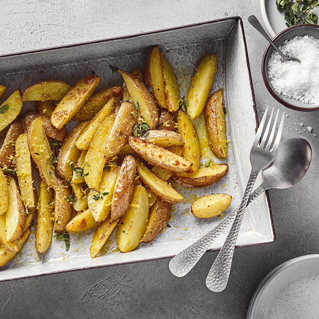 Ein Blech mit gebackenen Kartoffelspalten, bestreut mit gelben Zitronenzesten und grünen Kräutern.