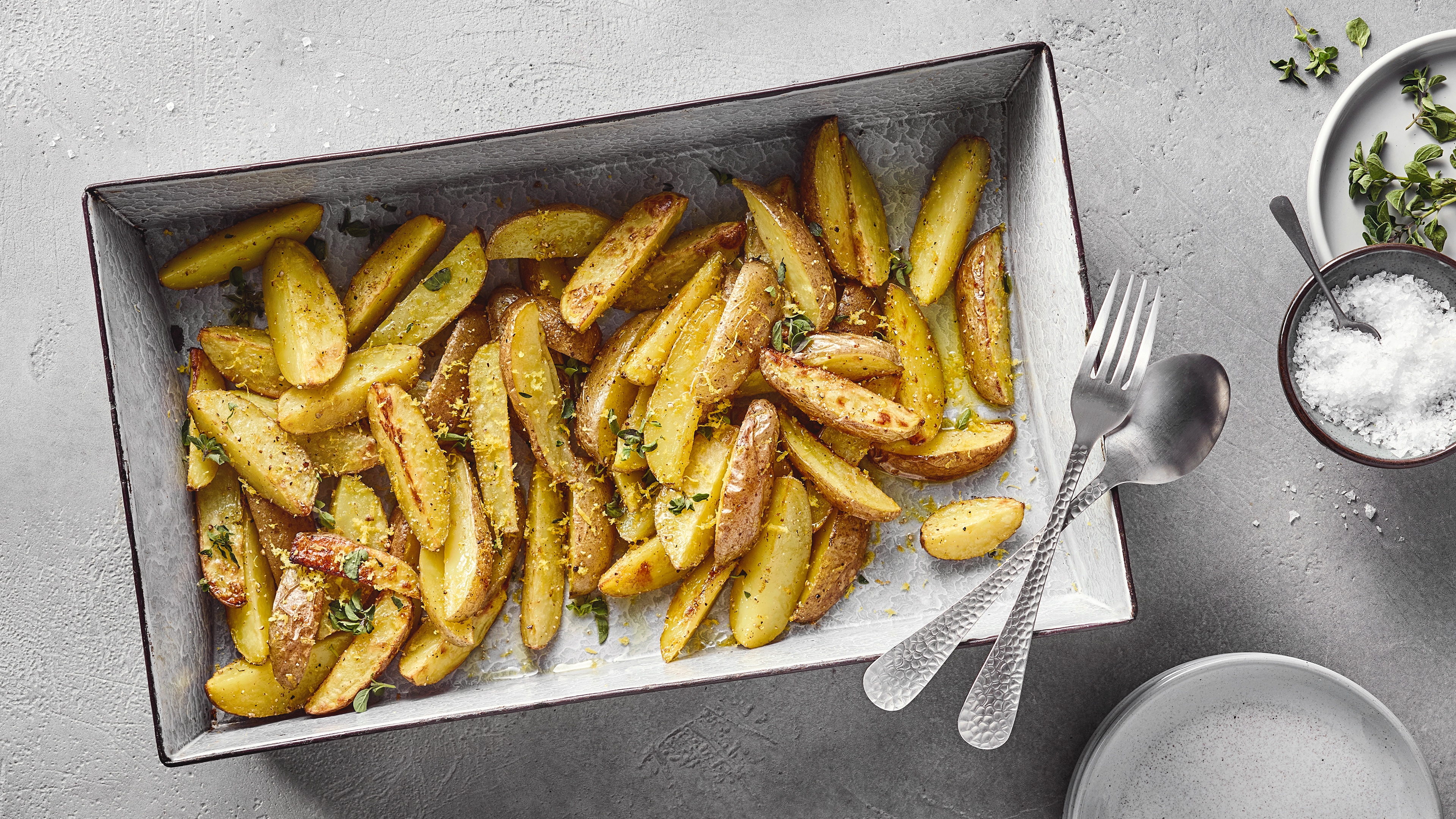 Ein Blech mit gebackenen Kartoffelspalten, bestreut mit gelben Zitronenzesten und grünen Kräutern.