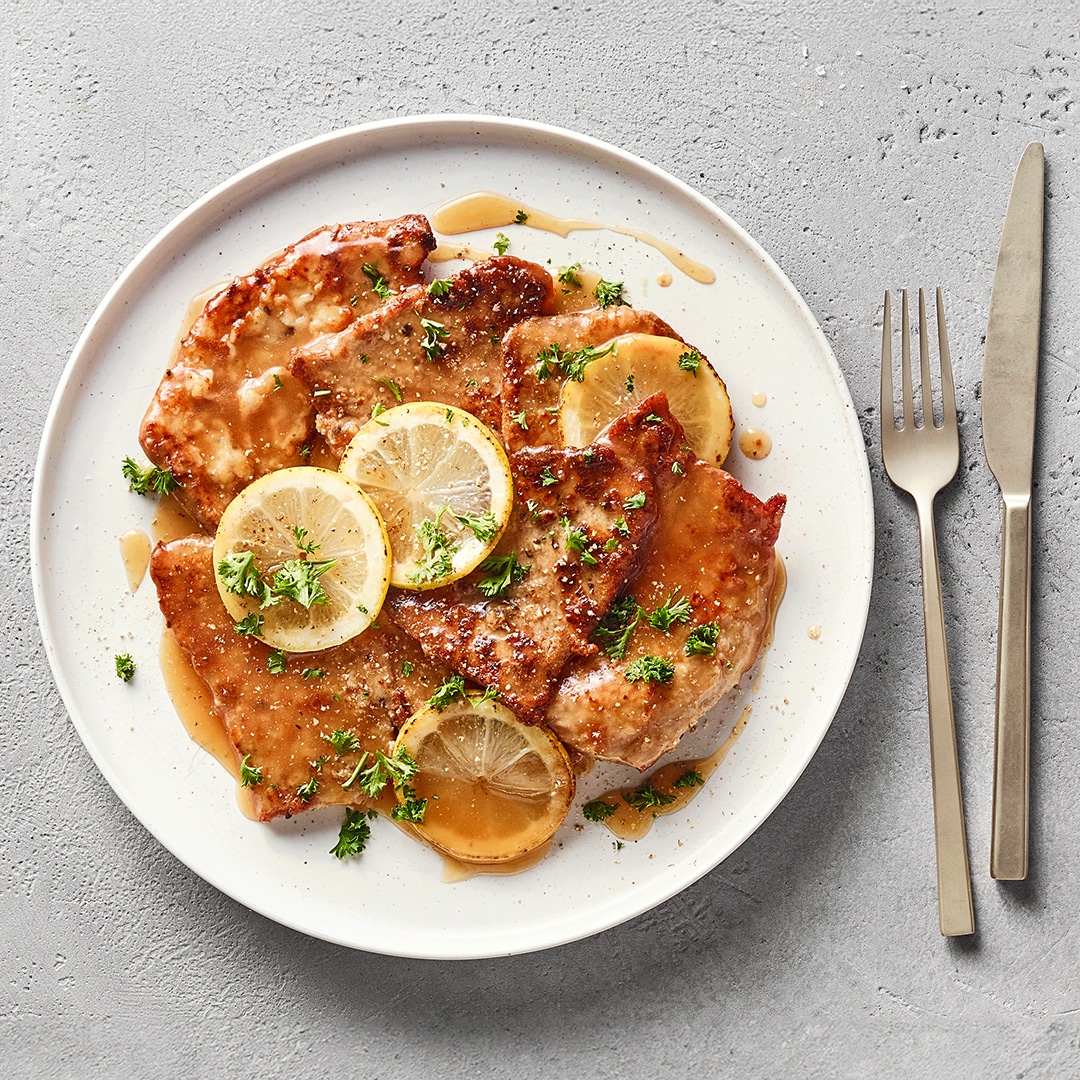 Ein Teller mit gebratenen Fleischscheiben, belegt mit Zitronenscheiben, grünen Kräutern und überzogen mit brauner Soße.