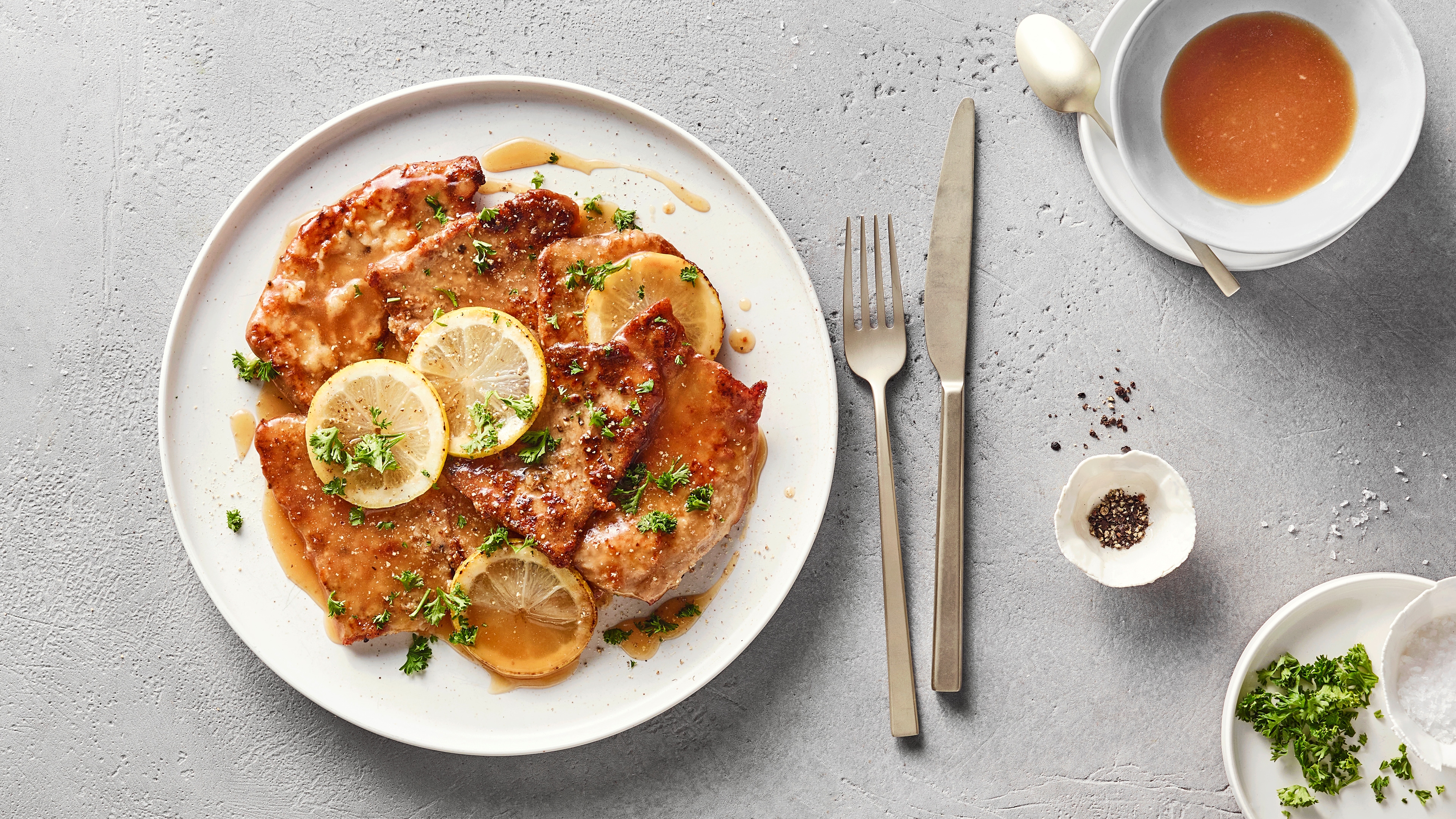 Ein Teller mit gebratenen Fleischscheiben, belegt mit Zitronenscheiben, grünen Kräutern und überzogen mit brauner Soße.