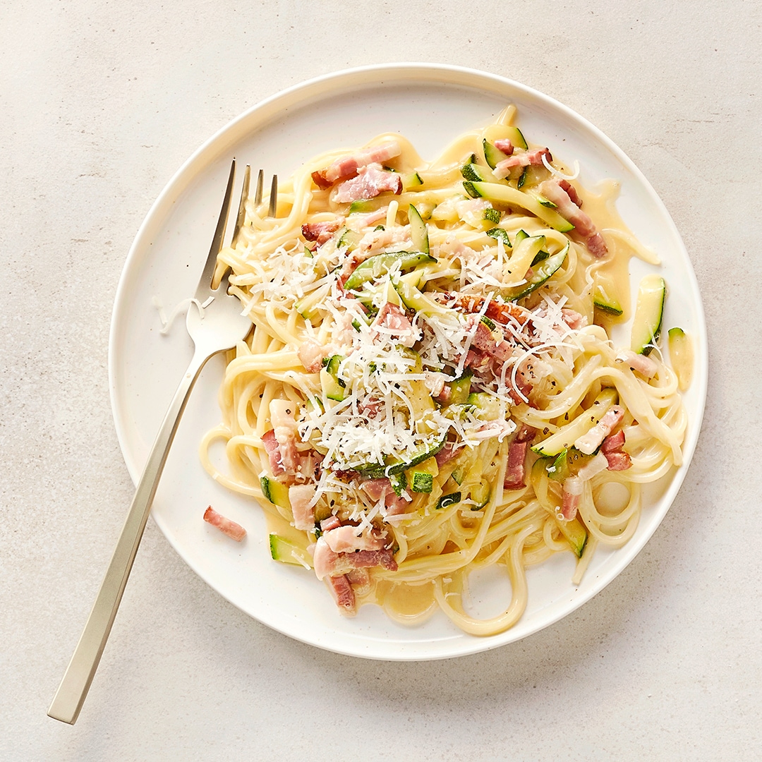 Ein Teller mit Spaghetti, Zucchinistreifen, gebratenem Speck und weißem geriebenem Käse in heller Soße.