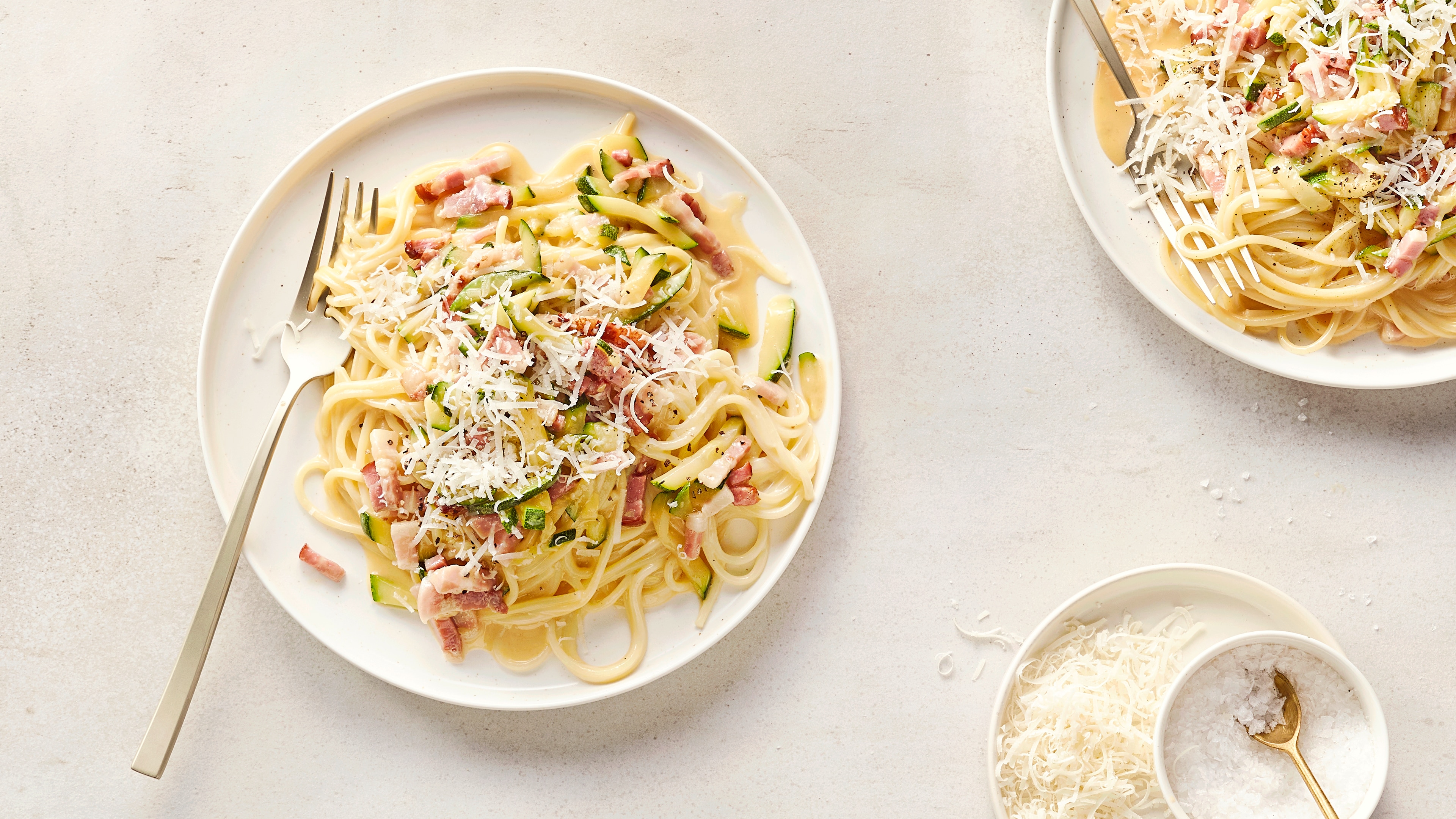 Ein Teller mit Spaghetti, Zucchinistreifen, gebratenem Speck und weißem geriebenem Käse in heller Soße.