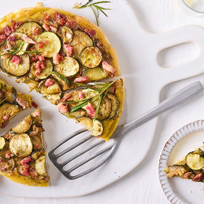 Eine große Tarte mit Zucchinischeiben, Speckwürfeln und Rosmarinzweigen