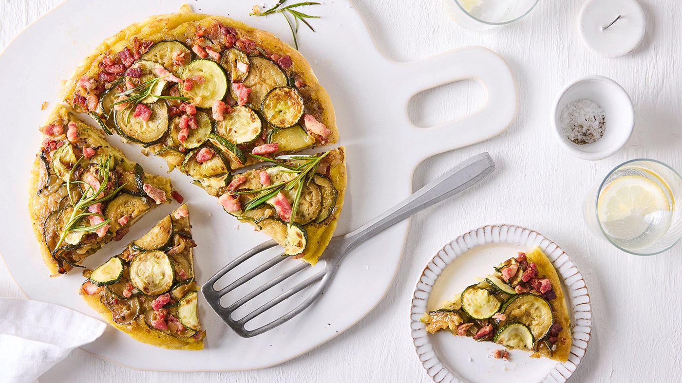 Eine große Tarte mit Zucchinischeiben, Speckwürfeln und Rosmarinzweigen. Daneben ein Teller mit einem Stück