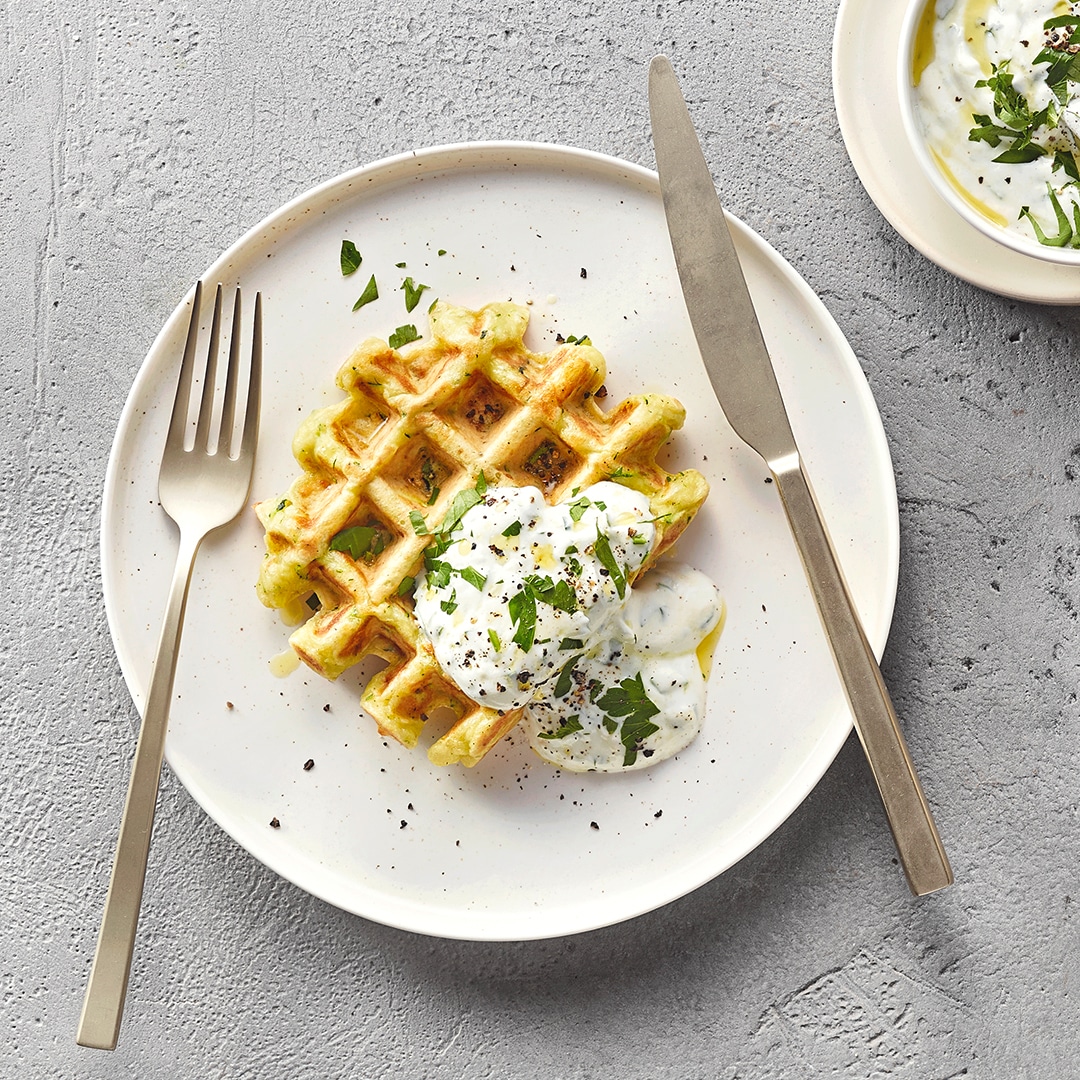 Ein Teller mit zwei goldbraunen Zucchiniwaffeln, darauf weißer Kräuterjoghurt und gehackte grüne Kräuter.