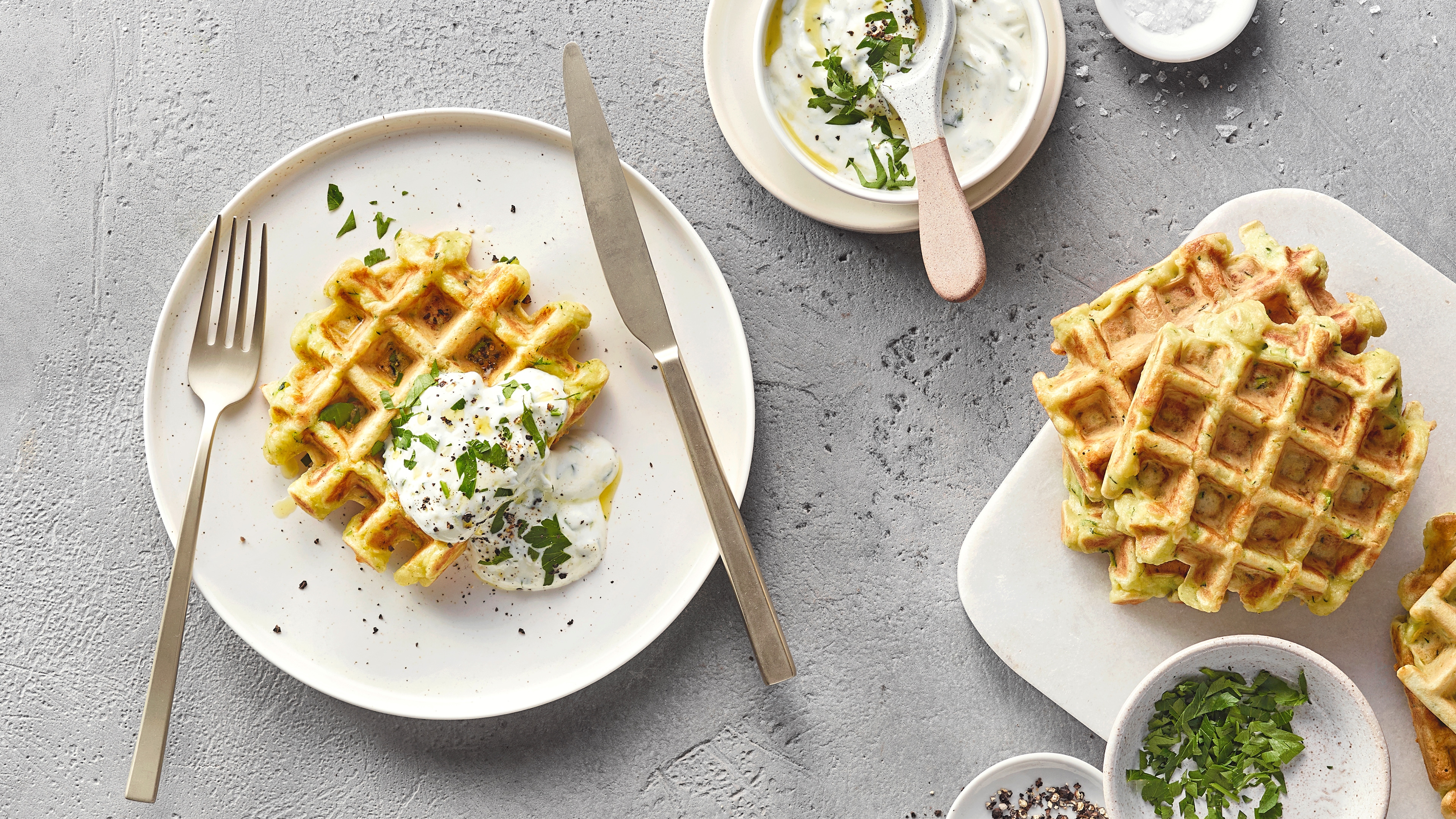 Ein Teller mit zwei goldbraunen Zucchiniwaffeln, darauf weißer Kräuterjoghurt und gehackte grüne Kräuter.
