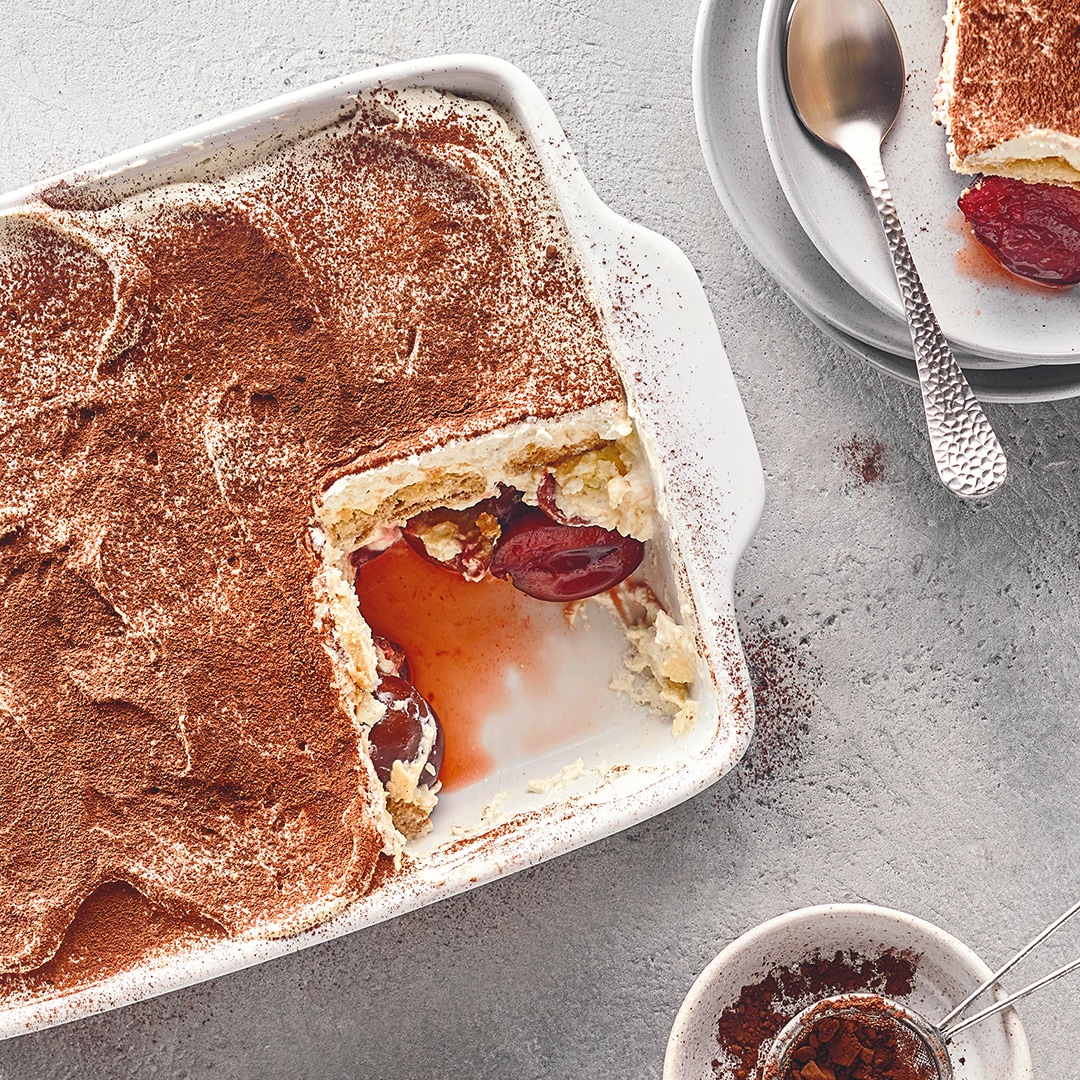 Auflaufform mit cremigem Schichtdessert aus heller Creme und Zwetschgenstücken, garniert mit Kakaopulver