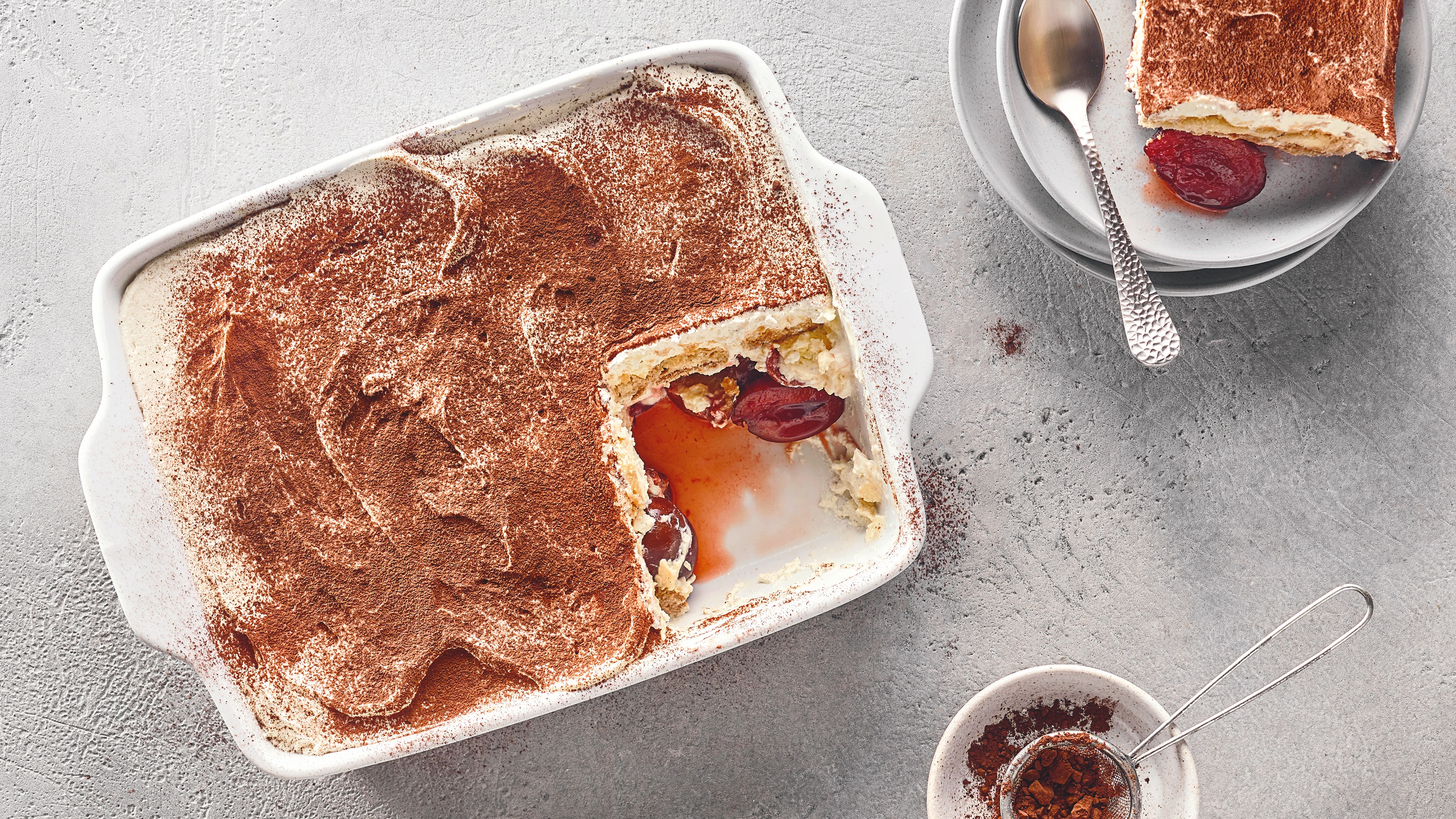 Auflaufform mit cremigem Schichtdessert aus heller Creme und Zwetschgenstücken, garniert mit Kakaopulver