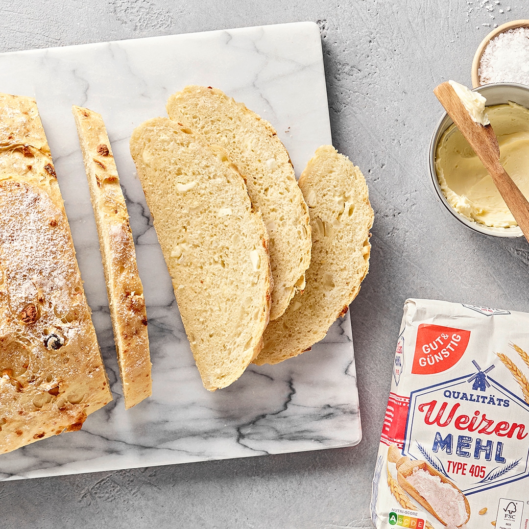 Angeschnittenes, rustikales Weißbrot mit Zwiebeln auf einer Marmorplatte, daneben GUT&GÜNSTIG Weizenmehl, Brotscheiben und ein Schälchen mit Butter.