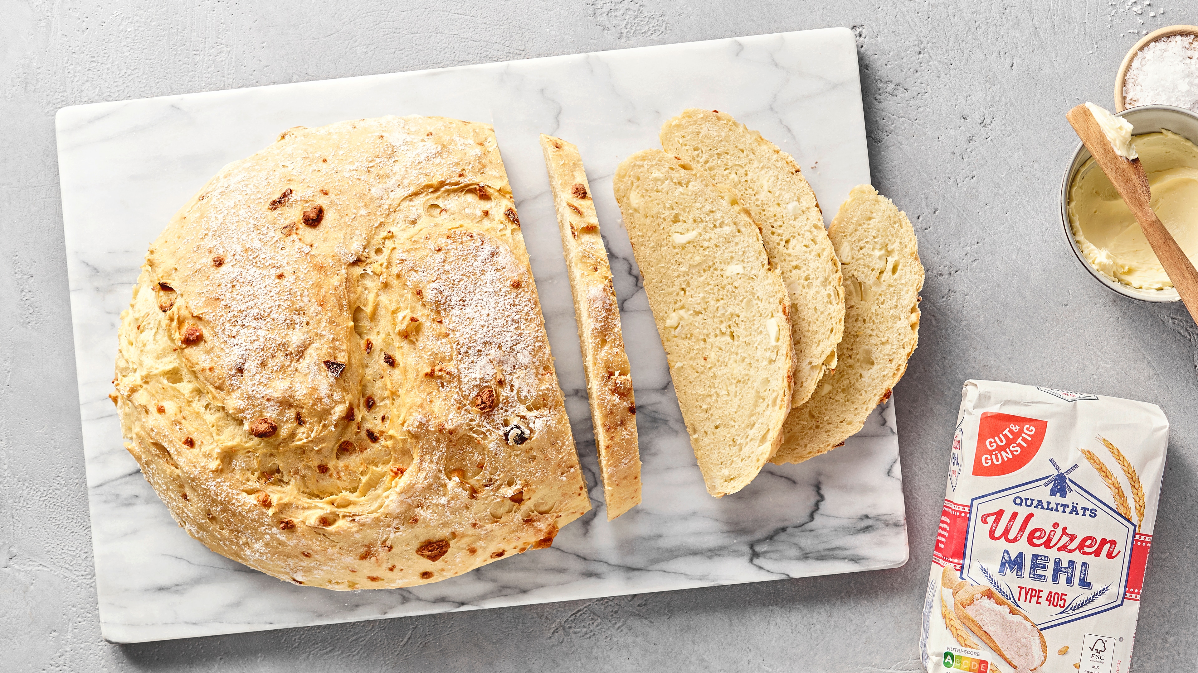 Angeschnittenes, rustikales Weißbrot mit Zwiebeln auf einer Marmorplatte, daneben GUT&GÜNSTIG Weizenmehl, Brotscheiben und ein Schälchen mit Butter.