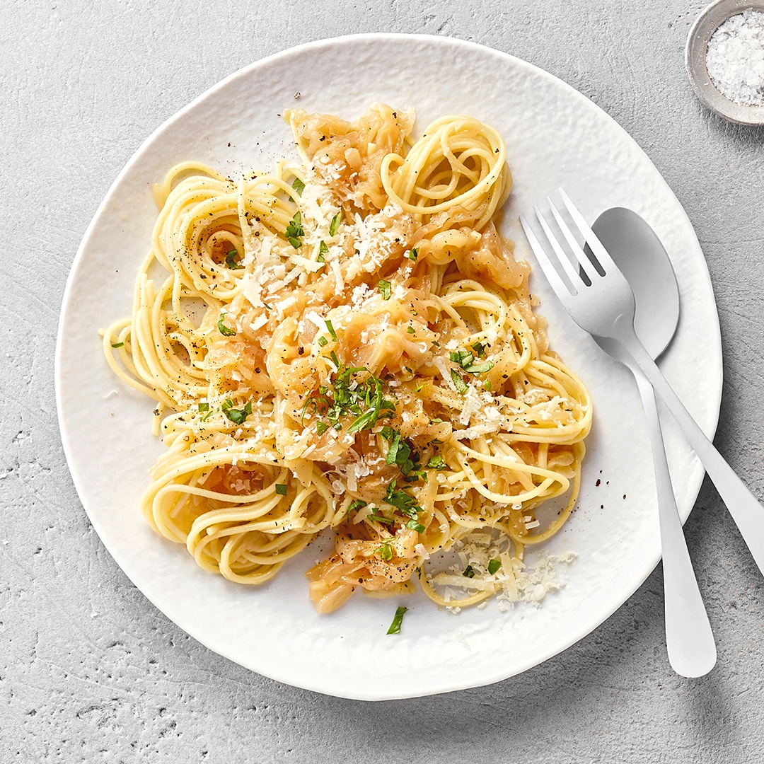 Spaghetti mit geriebenem Käse und Zwiebeln auf einem Teller, dazu Gewürze und Besteck.
