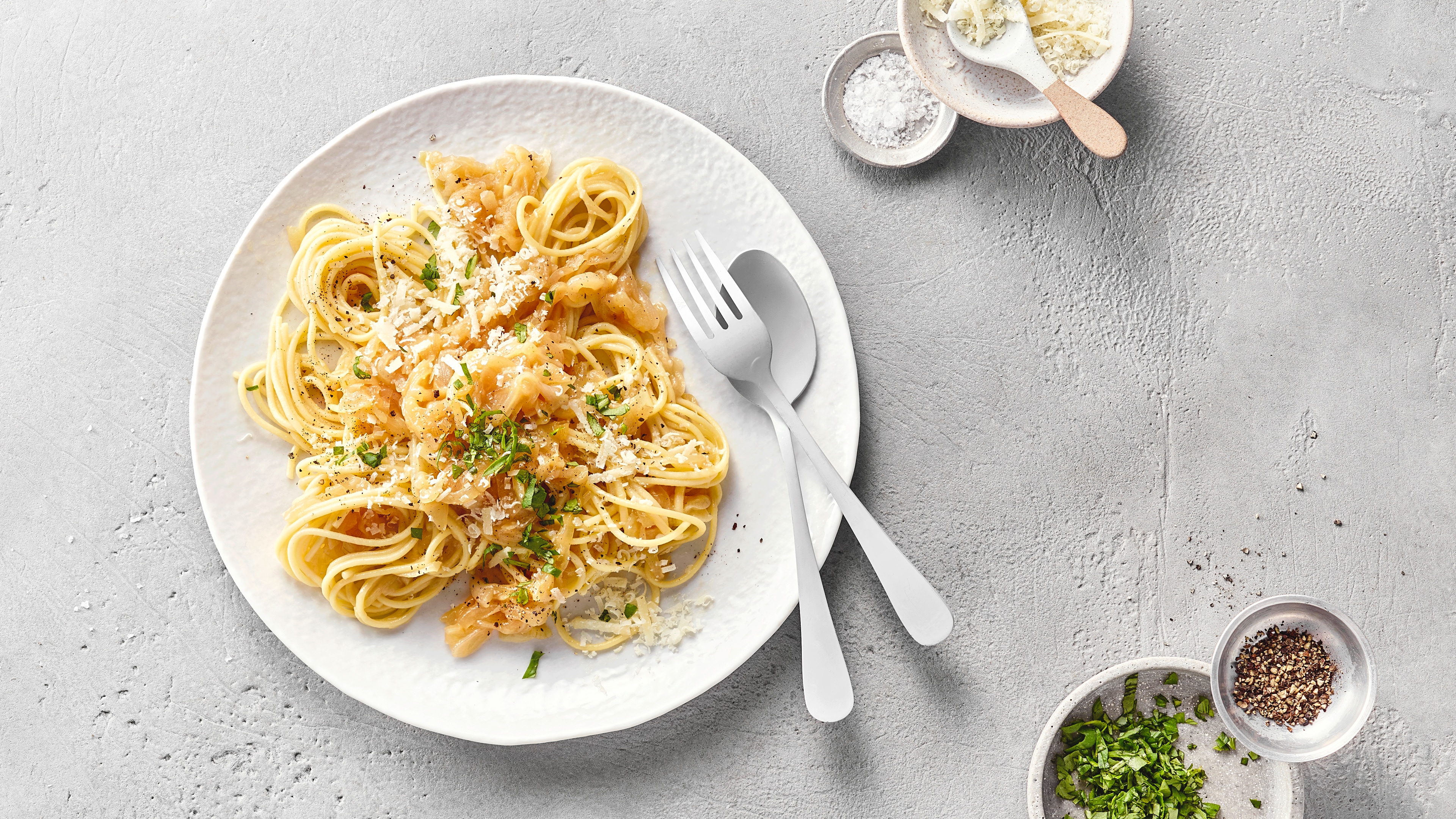 Spaghetti mit geriebenem Käse und Zwiebeln auf einem Teller, dazu Gewürze und Besteck.