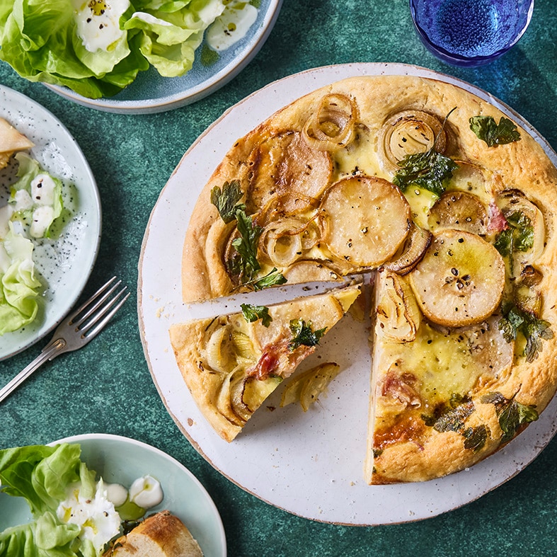Zwiebelkuchen mit Birnenscheiben und Pancetta auf einem Teller, dazu grüner Salat und gedeckter Tisch.