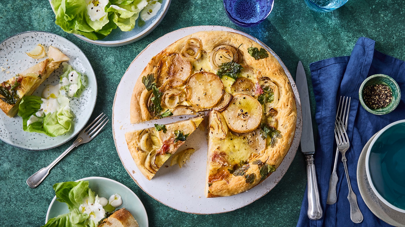 Zwiebelkuchen mit Birnenscheiben und Pancetta auf einem Teller, dazu grüner Salat und gedeckter Tisch.